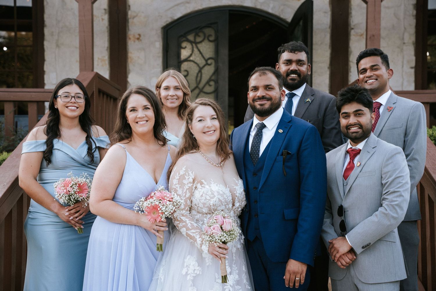 A wedding party in blue and pink attire poses together outside a brick building.