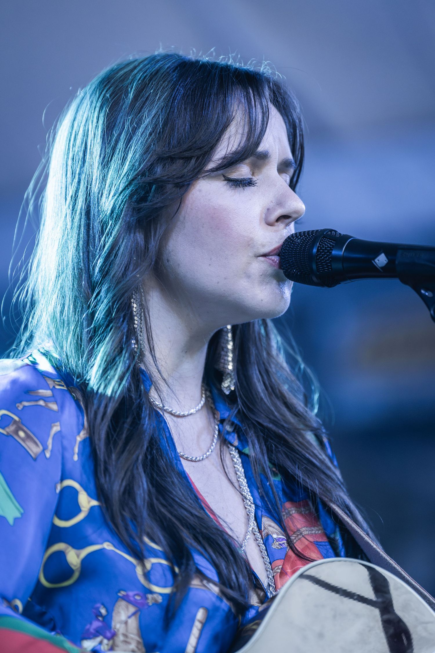 Singer performs on stage wearing a vibrant blue patterned shirt while singing into a microphone in atmospheric lighting.