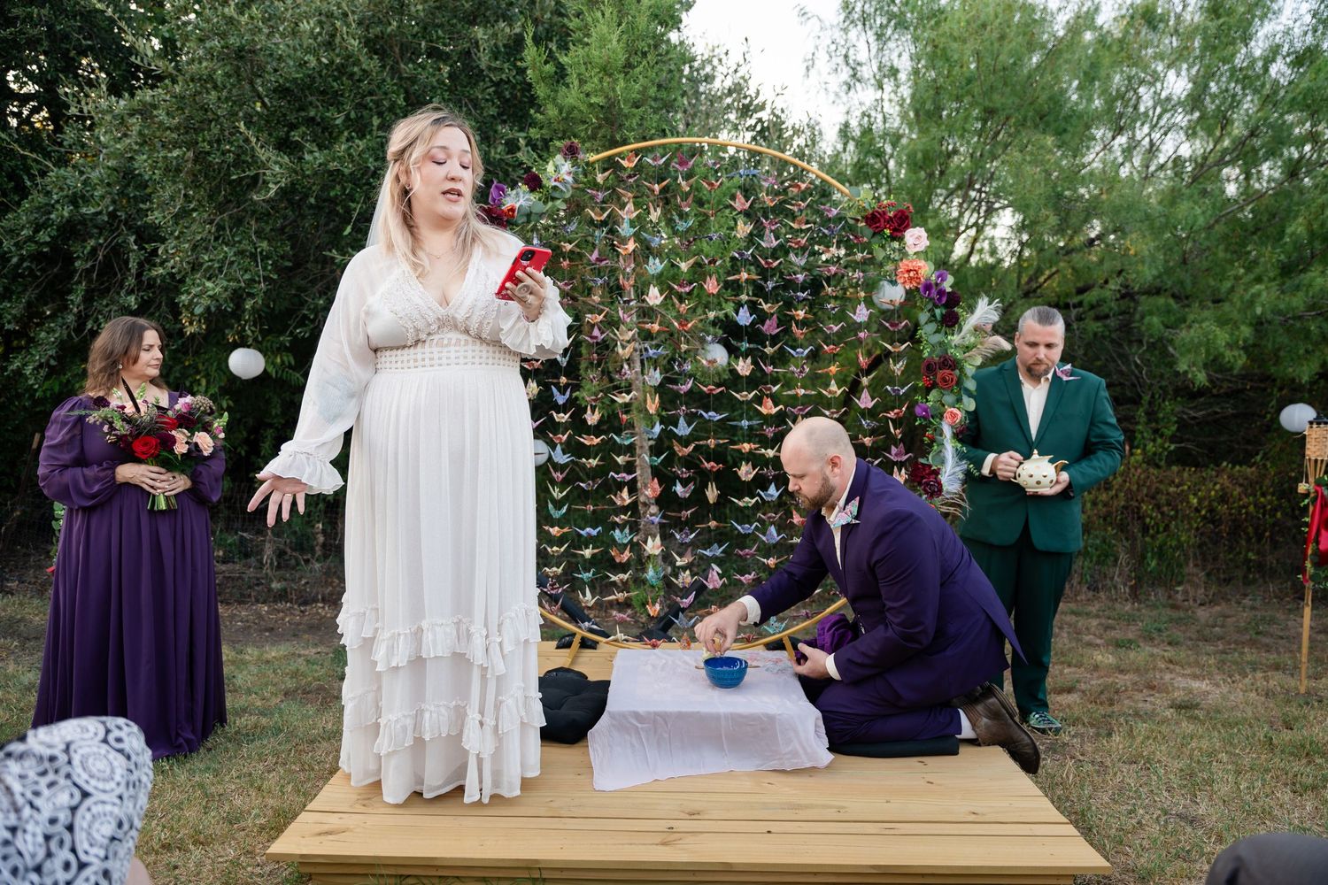 Wedding couple participates in ceremony ritual before paper crane backdrop in garden setting.