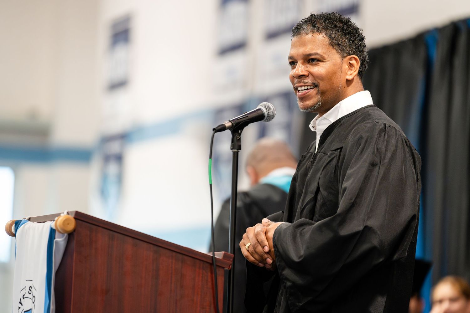 Speaker in black graduation robe delivers speech from podium during commencement ceremony.