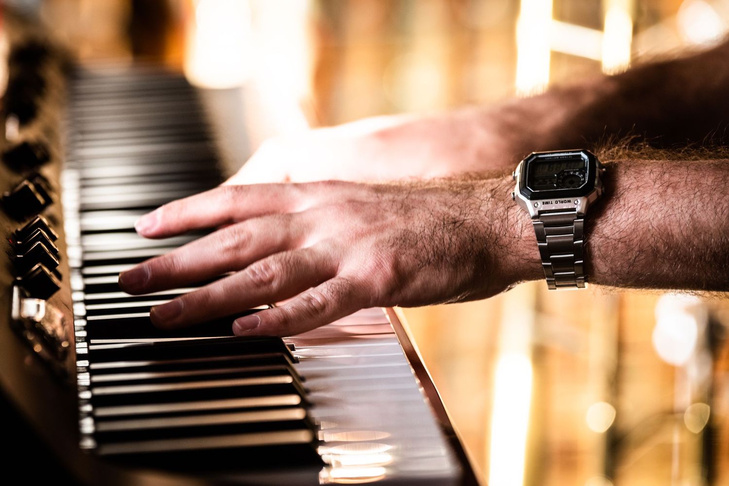 Close-up of hands wearing digital watch playing piano keys in warm lighting.