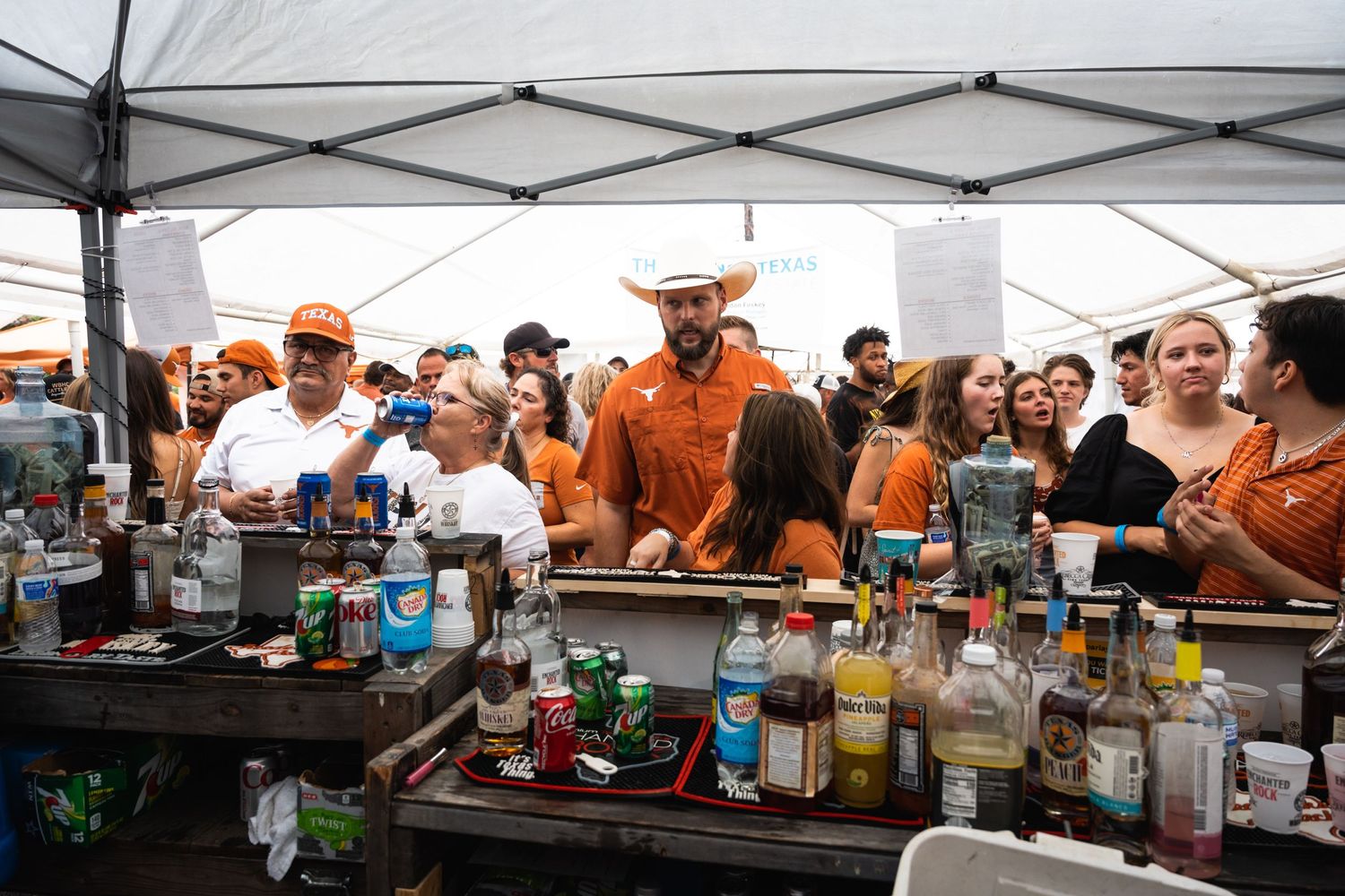 Crowd gathers around outdoor bar setup with multiple bottles and servers.