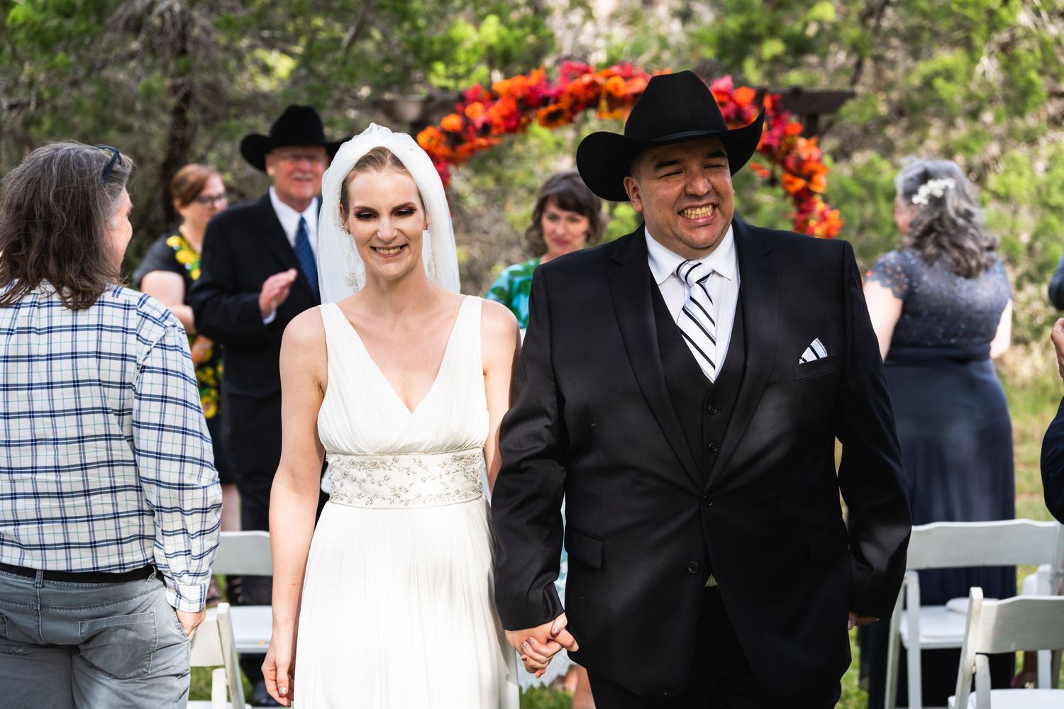 Newlyweds walk hand in hand past wedding guests under a bright floral arch.