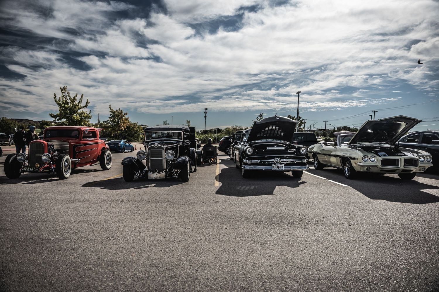 Vintage hot rods and classic cars lined up at outdoor auto show under cloudy skies.
