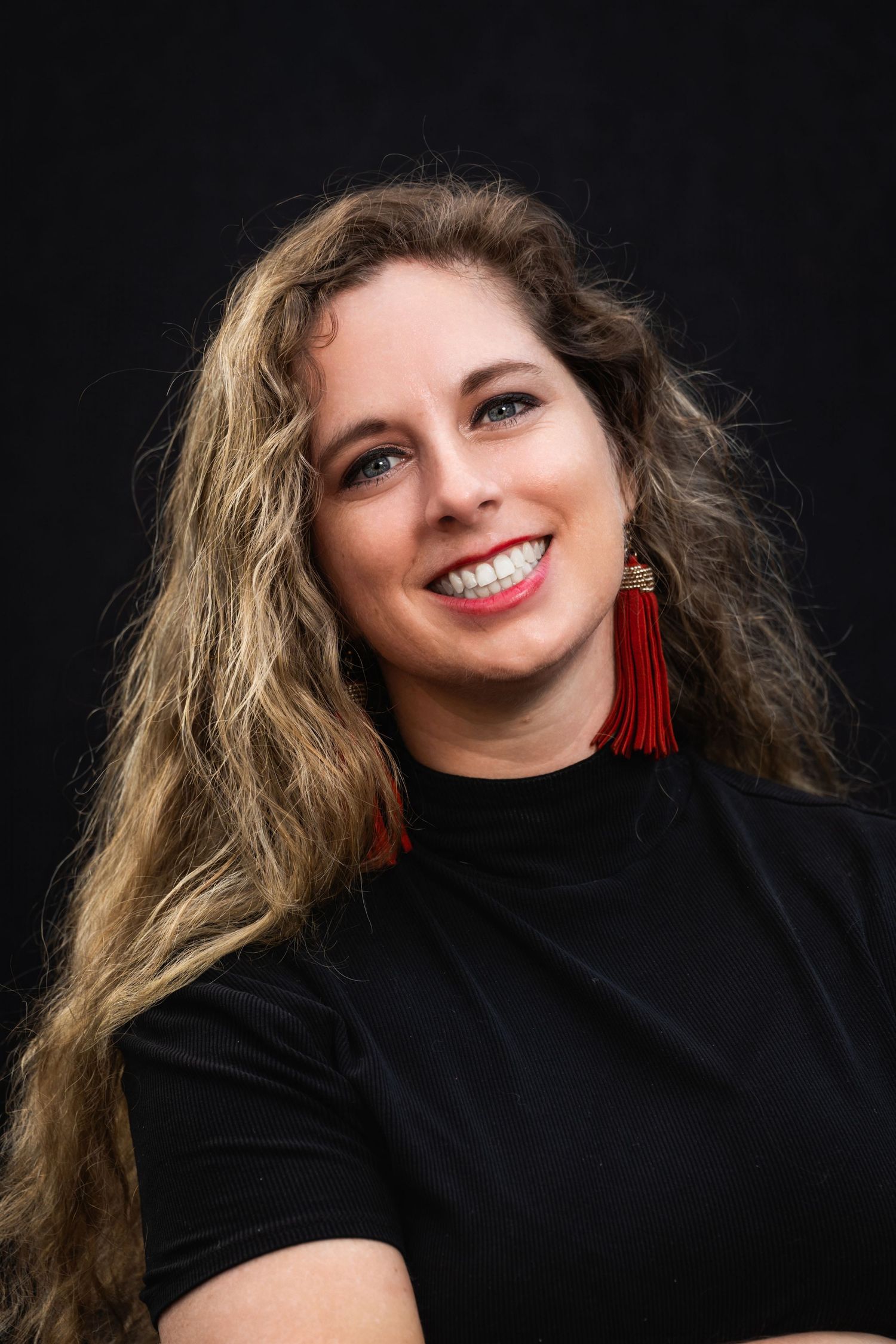 Close-up portrait of woman in black top with red tassel earrings against dark background.