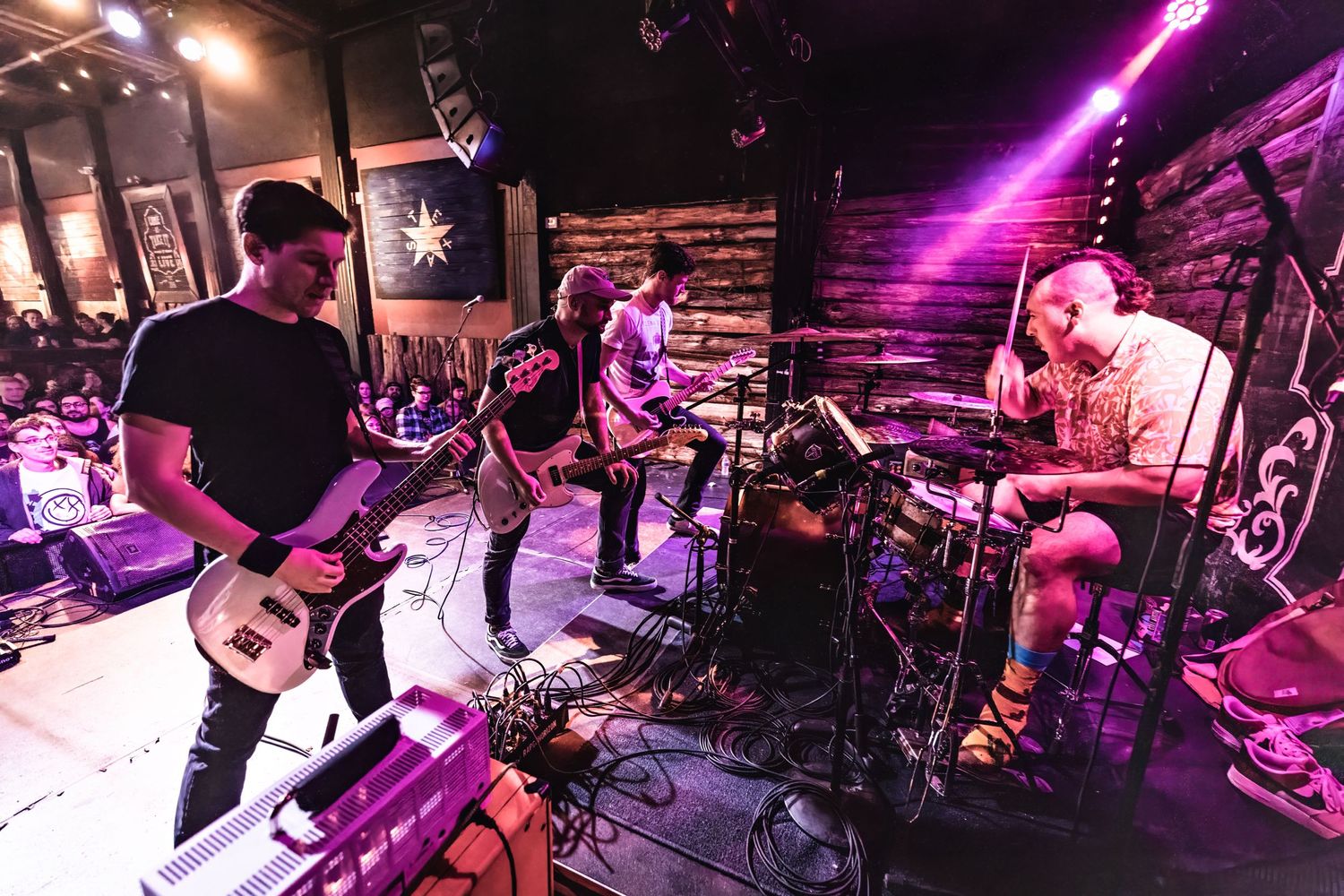 Rock band performs on stage with purple lighting and wooden wall backdrop.