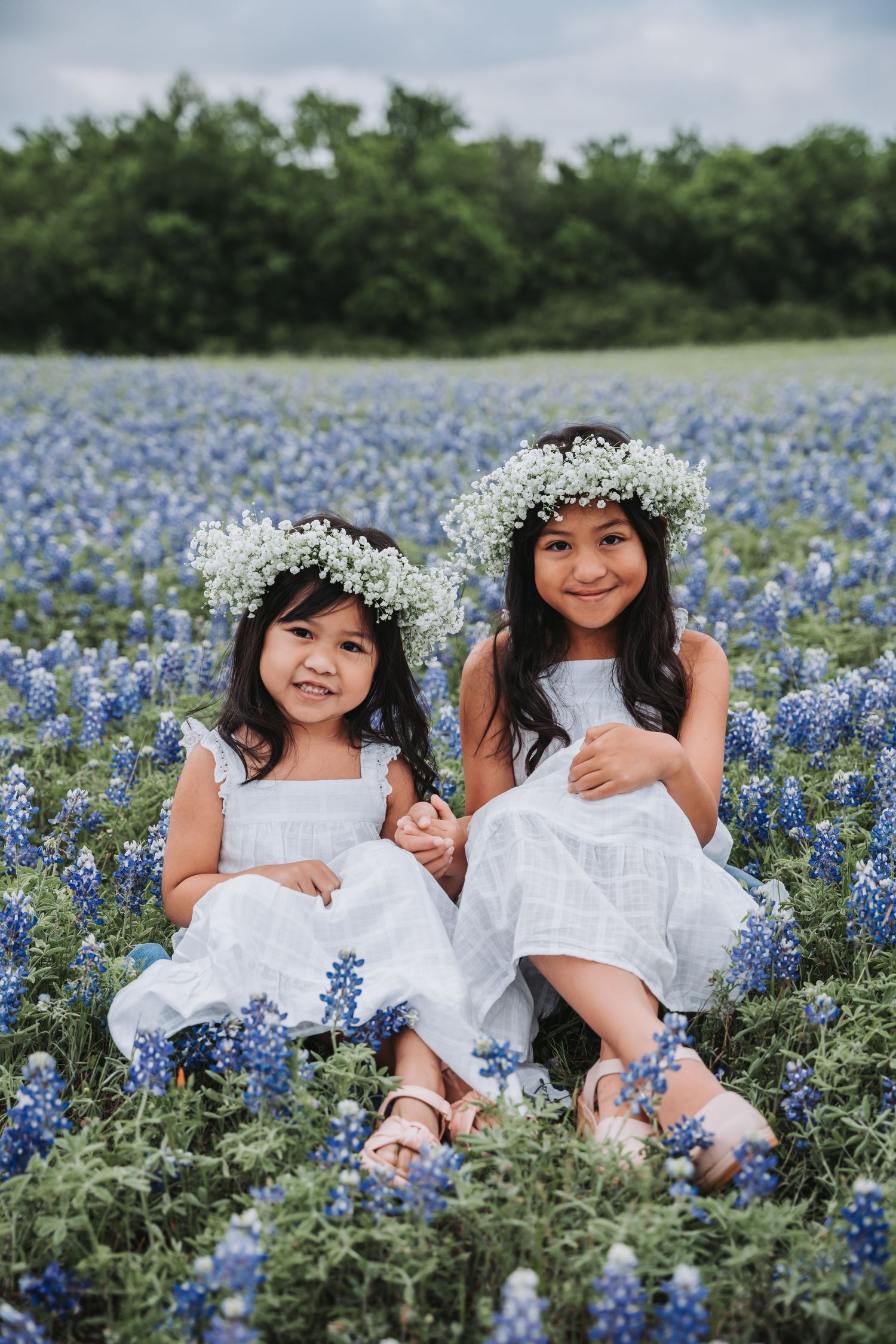 Two children wearing white dresses and floral crowns sit among vibrant bluebonnets in a spring meadow.