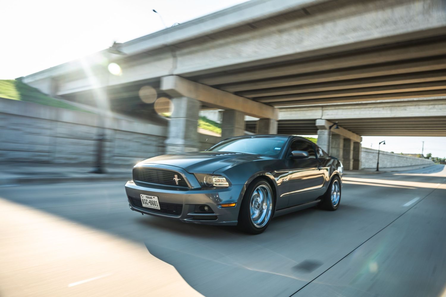 Sleek blue Mustang in motion beneath highway overpass with sunflare.