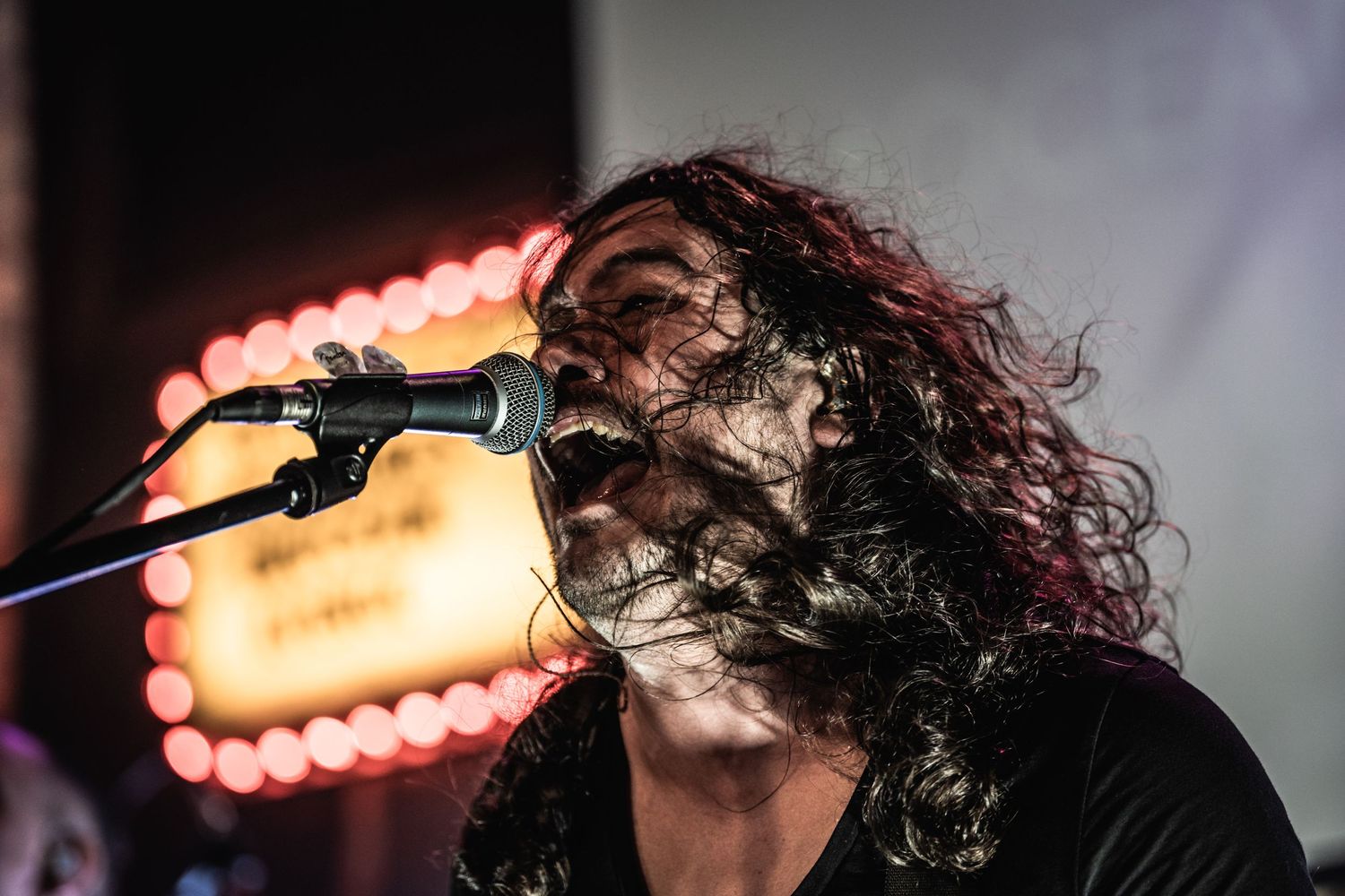 Concert performer with curly hair singing passionately into microphone with illuminated sign behind.