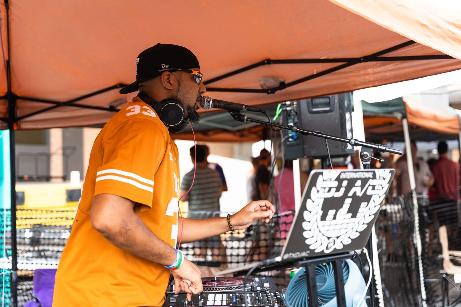 DJ in orange shirt performs at outdoor event under tent.