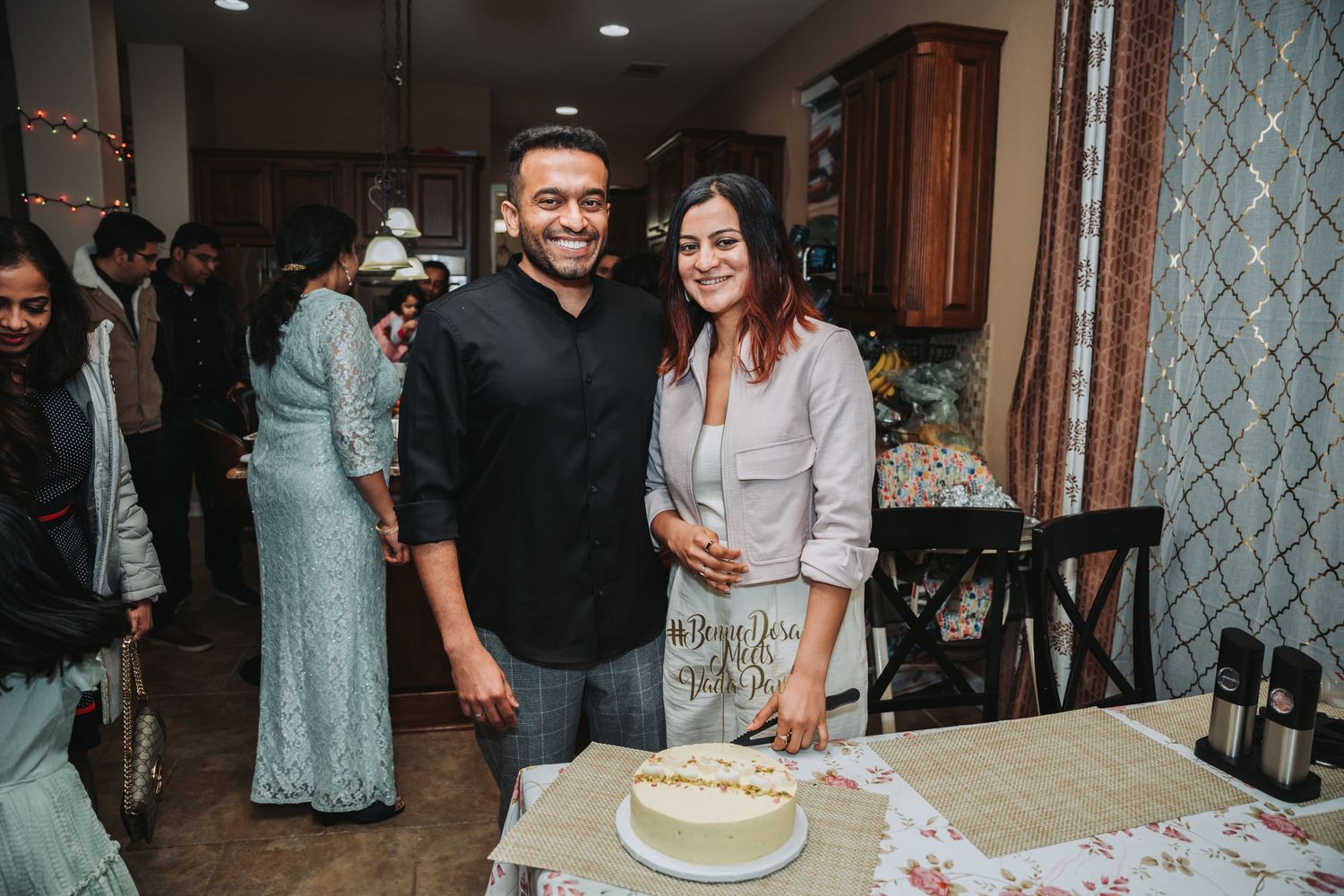 A couple poses together behind their white celebration cake at an indoor gathering.