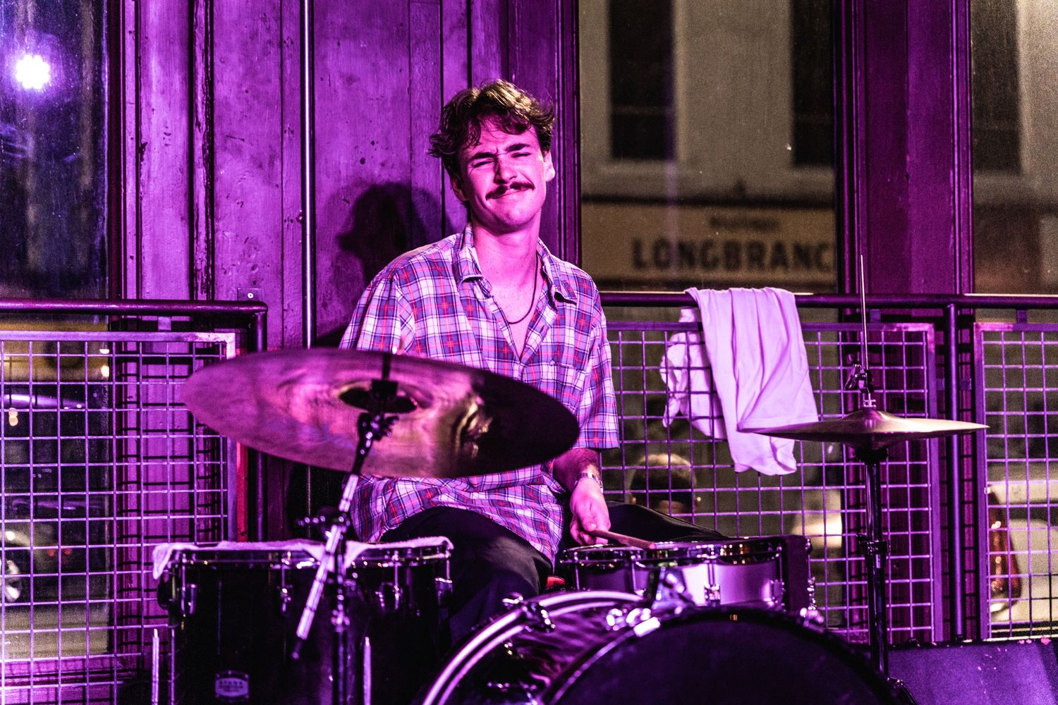 Drummer performing on stage bathed in purple lighting at music venue.