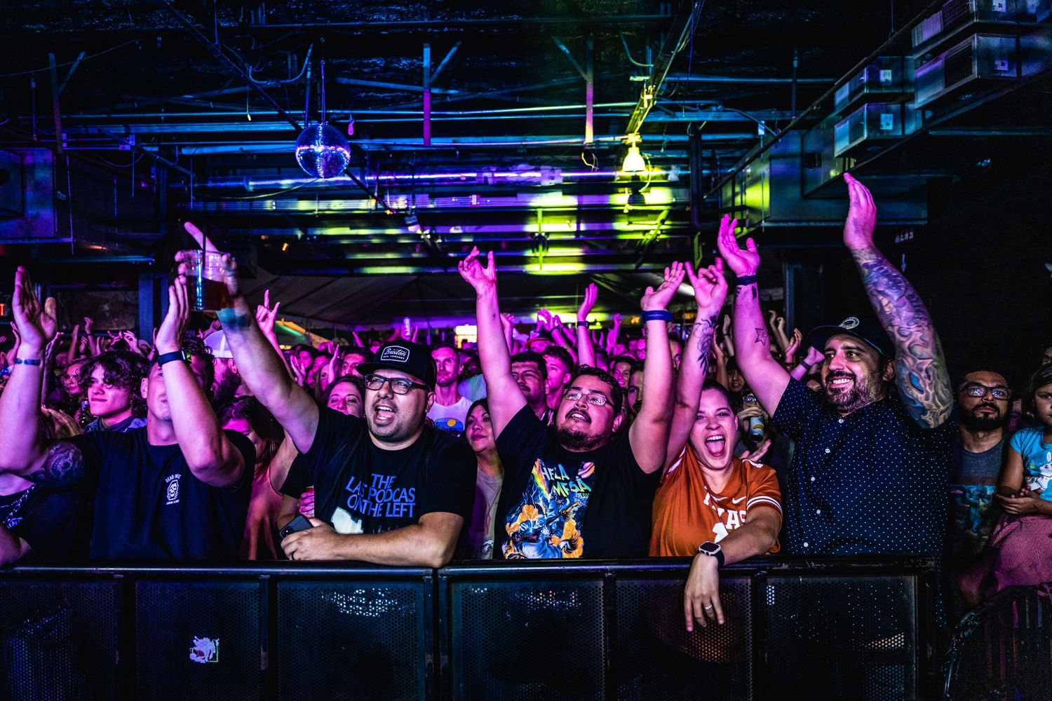 Excited concert crowd raises hands in unison under colorful venue lighting.