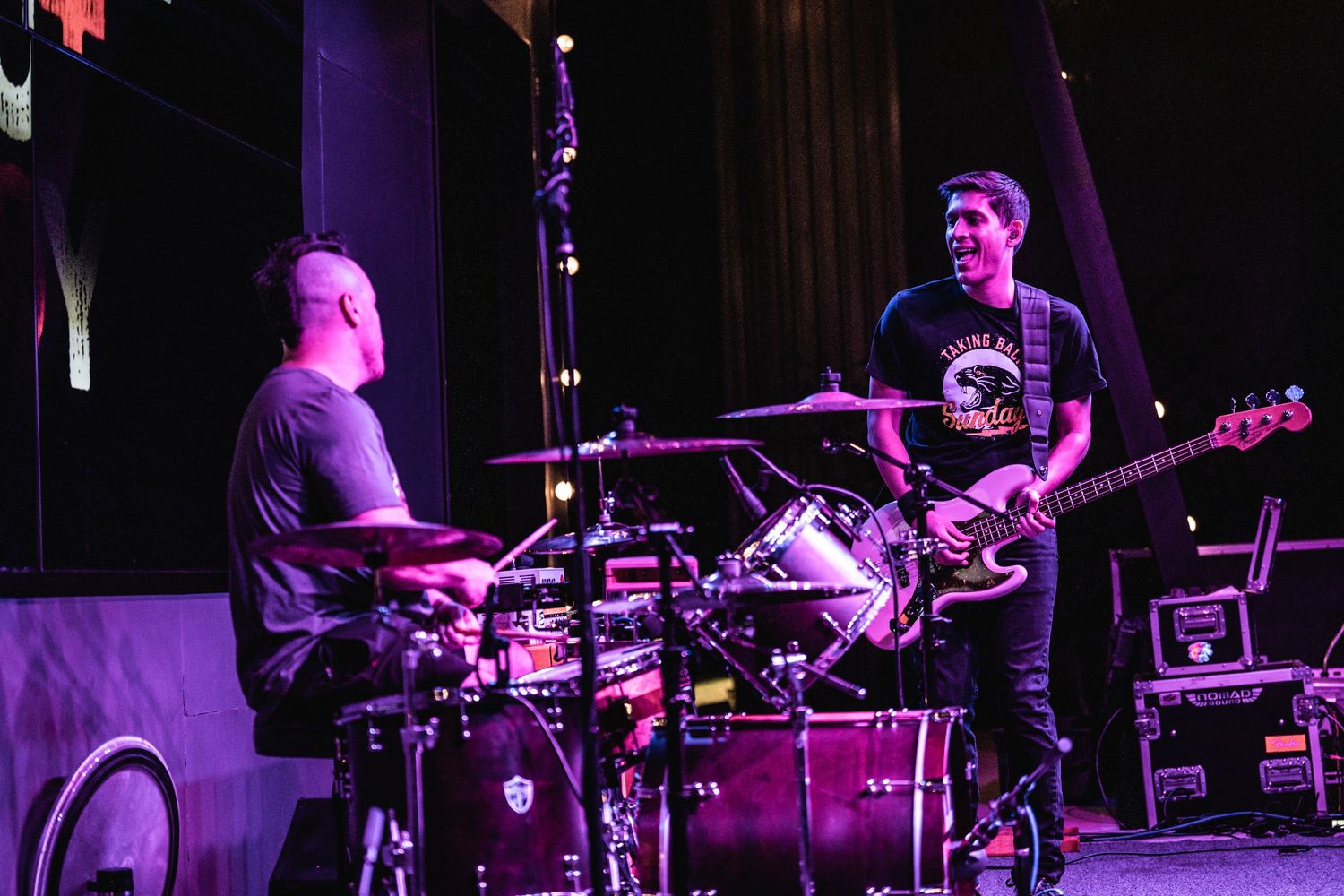 Musicians performing on purple-lit concert stage with drum kit visible.