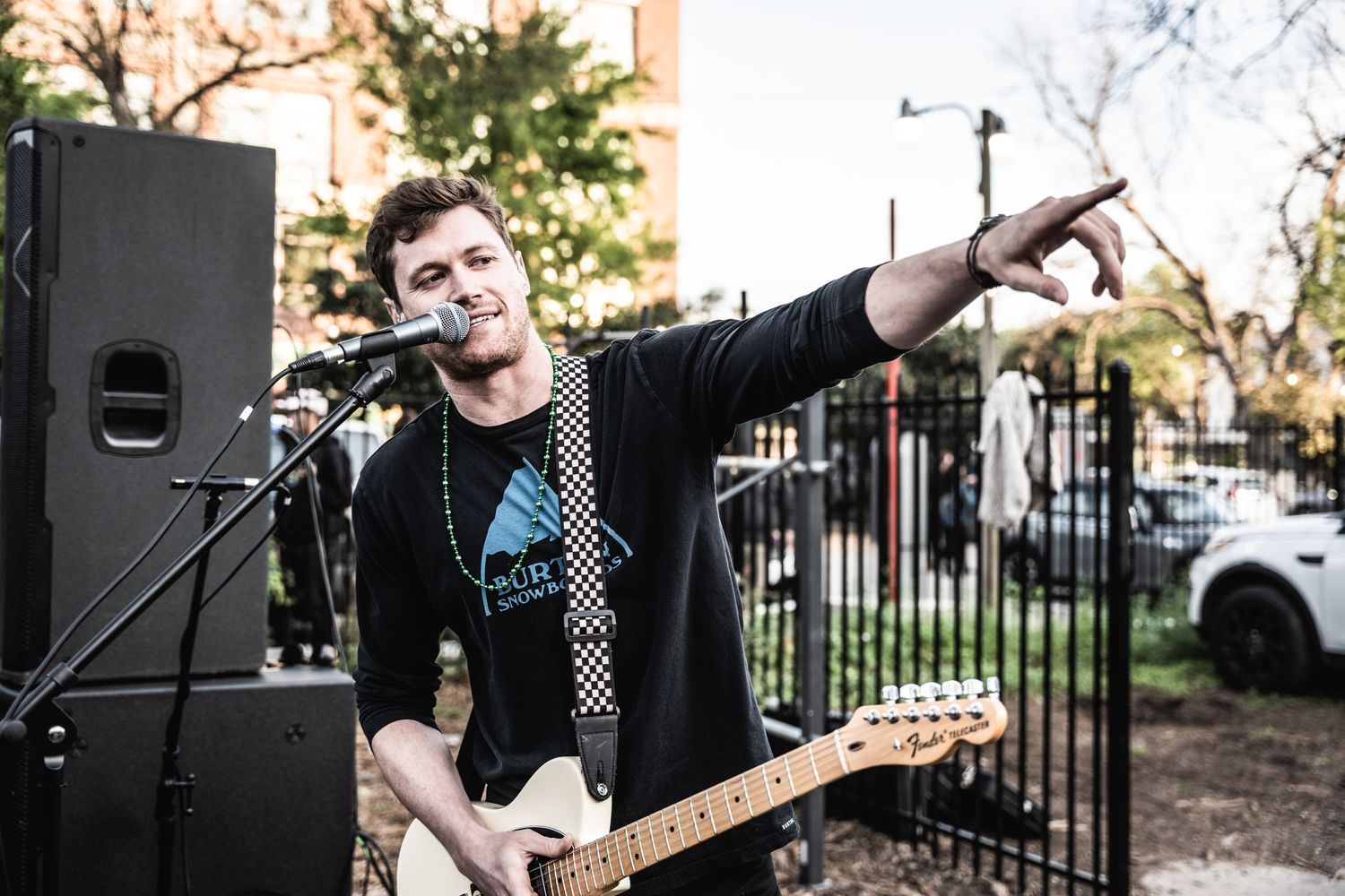 Performer with electric guitar gesturing while singing into microphone outdoors.