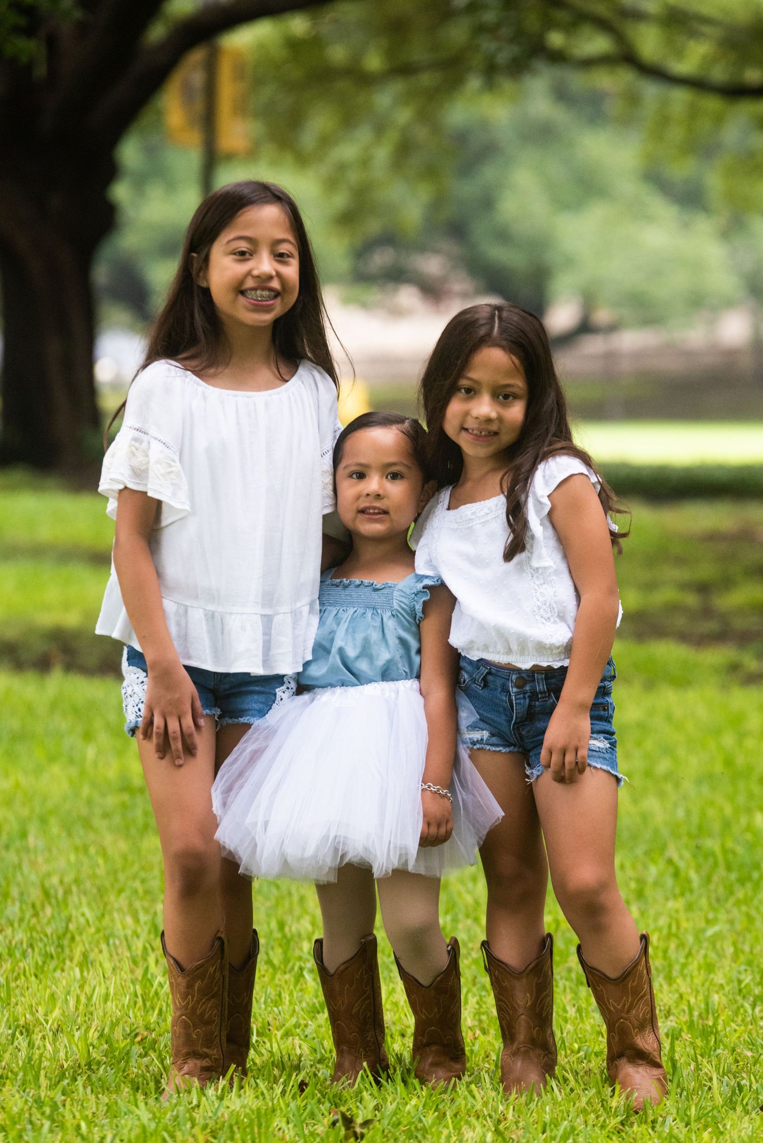 Three sisters in matching outfits with cowboy boots pose together on grass.