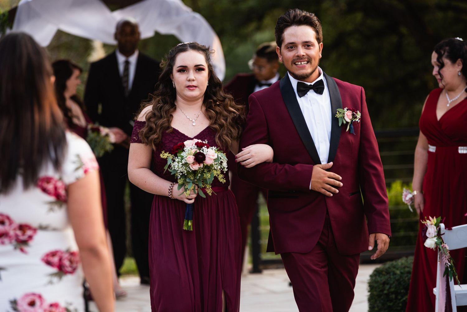 Wedding party members wearing burgundy formal attire walking during the ceremony.