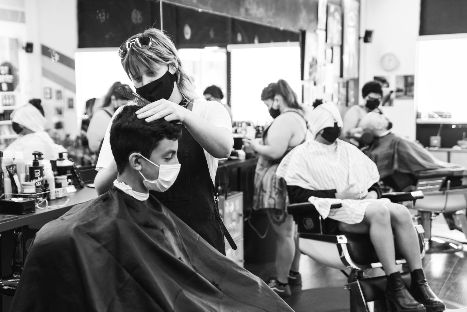 Black and white image of a busy barbershop interior with clients getting haircuts.