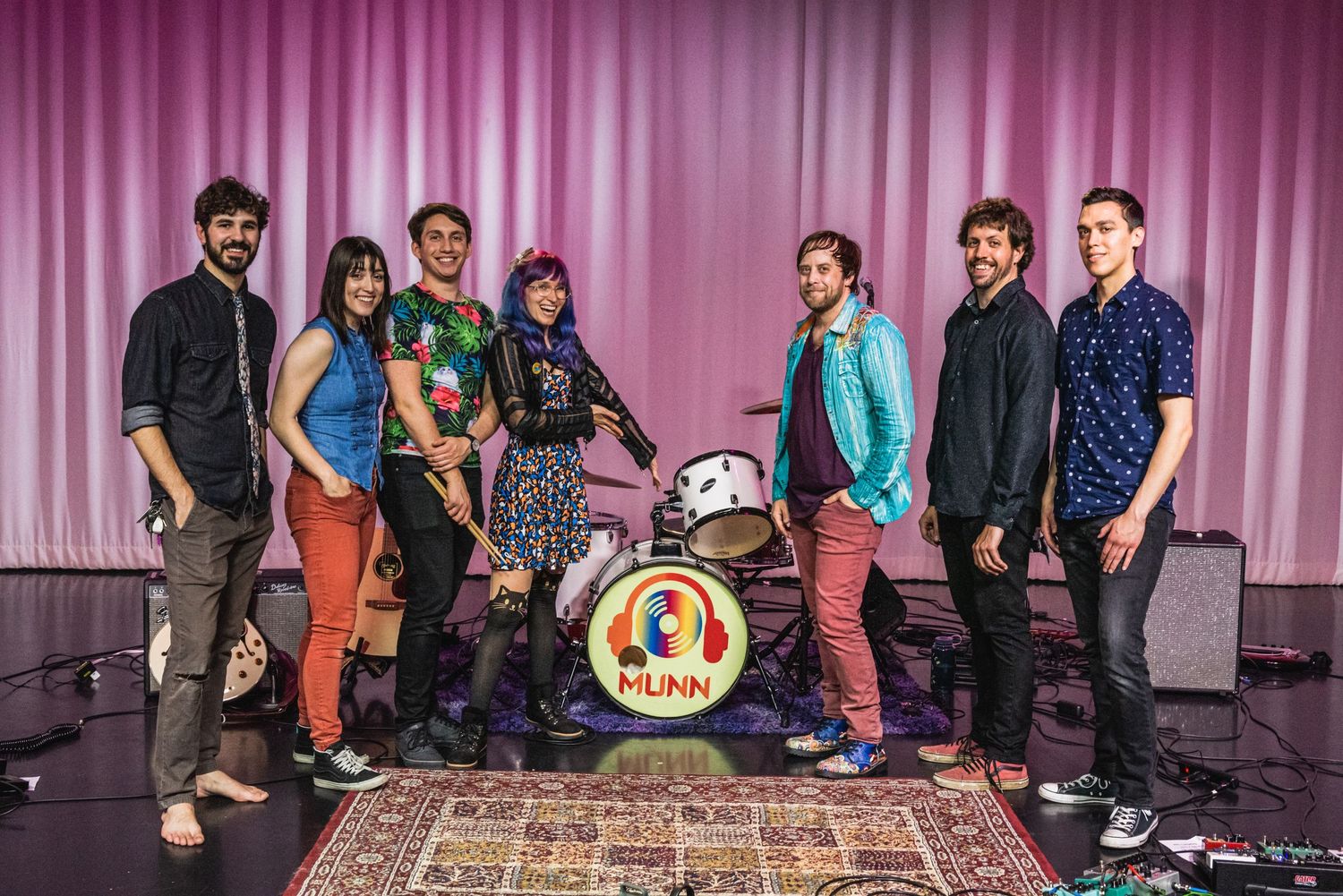 Band members pose together with their instruments on oriental rug against pink backdrop.