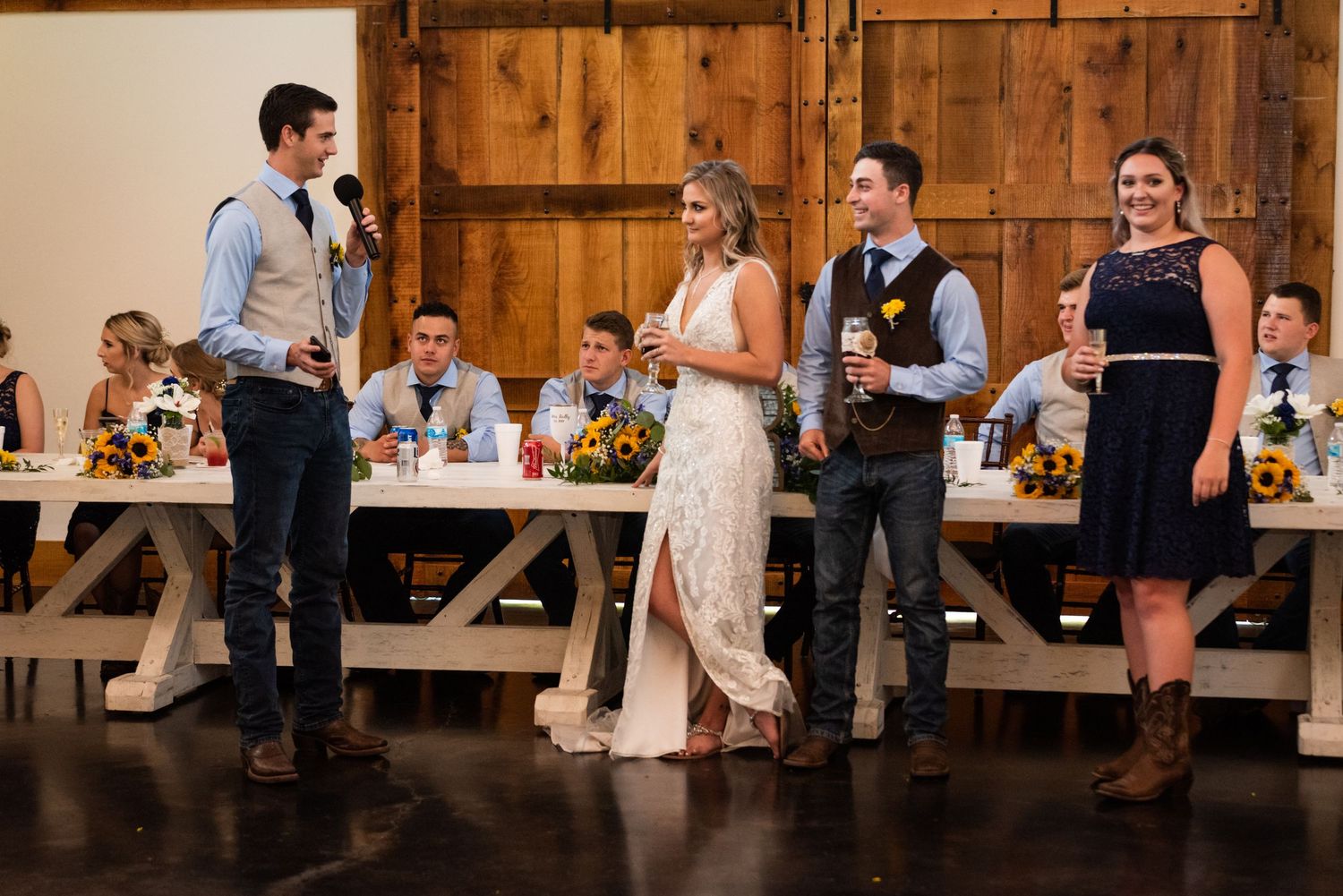 Wedding party members gathered for speeches in front of rustic barn doors at reception venue.