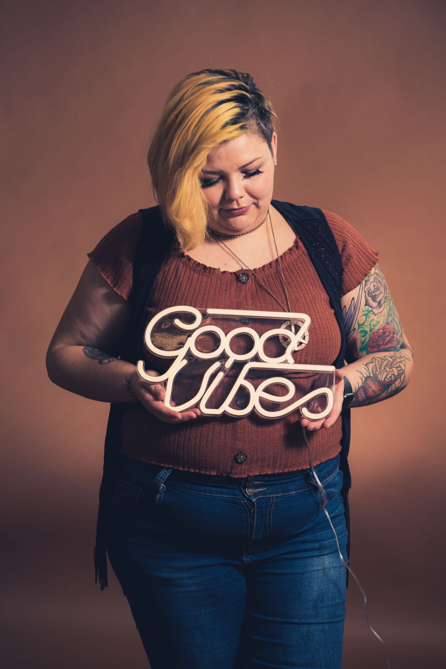 Person holding illuminated Good Vibes neon sign wearing brown top and black vest.