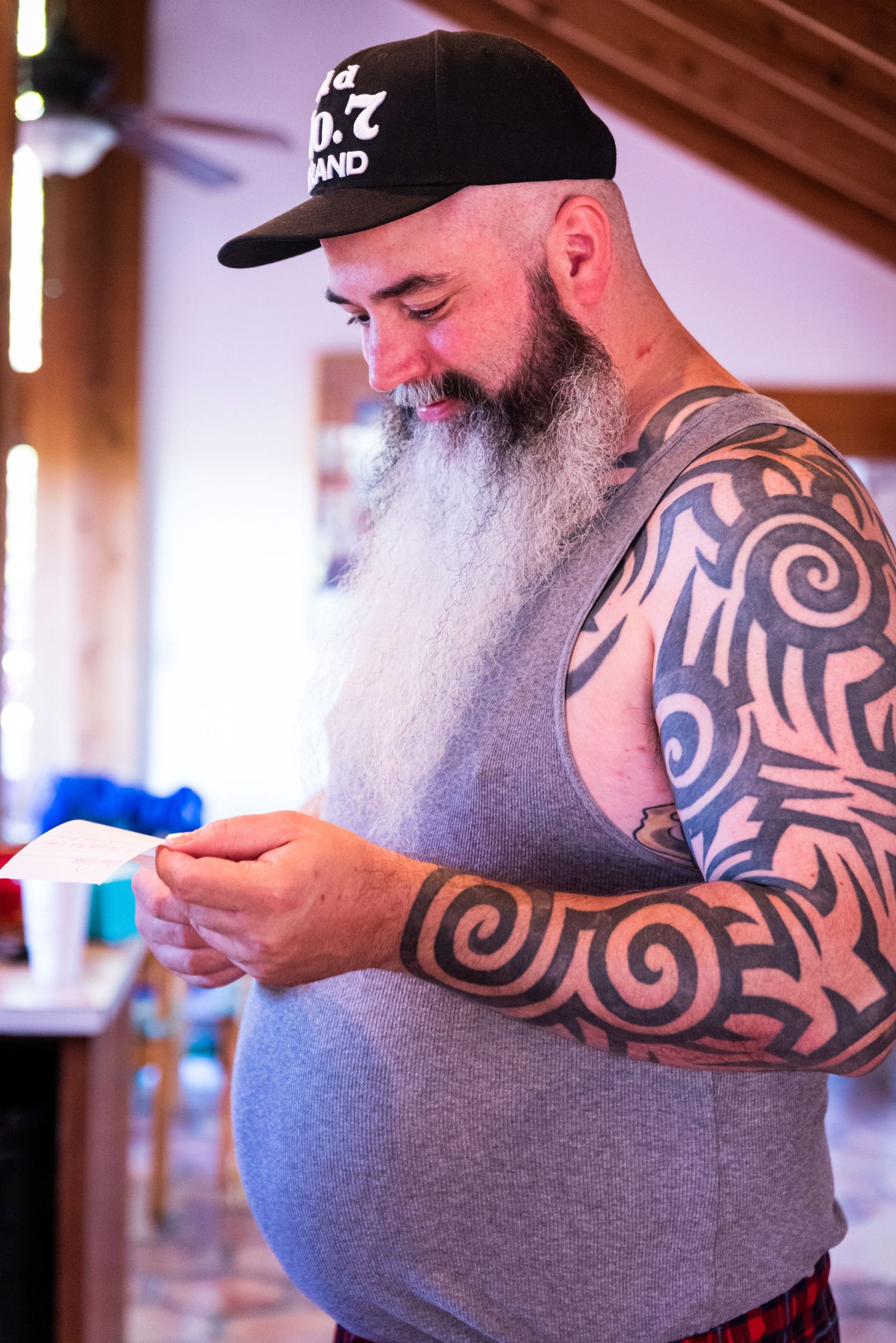 A bearded man with tribal tattoos wearing a black cap examines something in his hands.