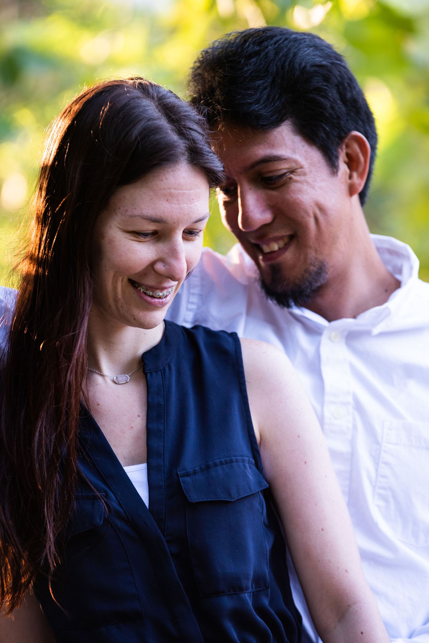 A happy couple share an intimate moment while laughing together outdoors with green foliage in the background.
