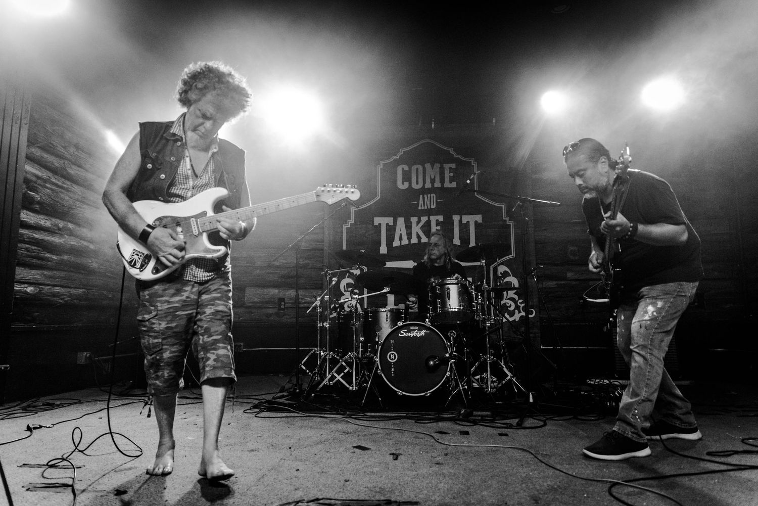 Black and white photo of band performing with drums and guitar players on small stage.