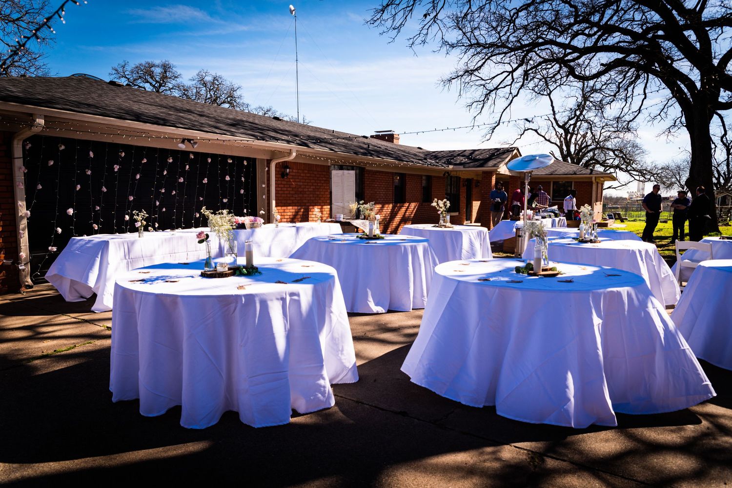 Outdoor wedding reception setup with white tablecloths and string lights under trees.