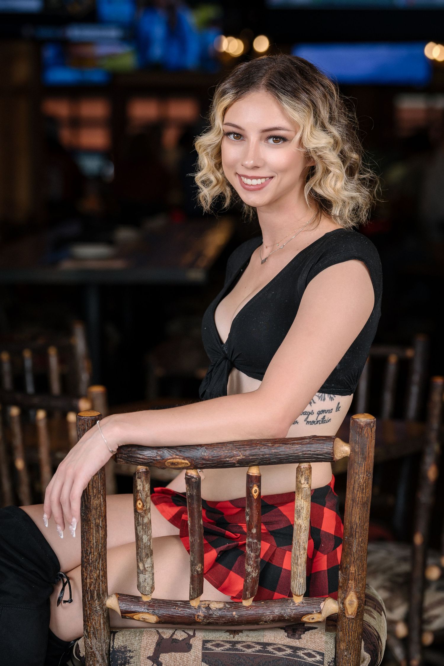 Server wearing casual black top and plaid skirt sits in rustic wooden chair at restaurant.