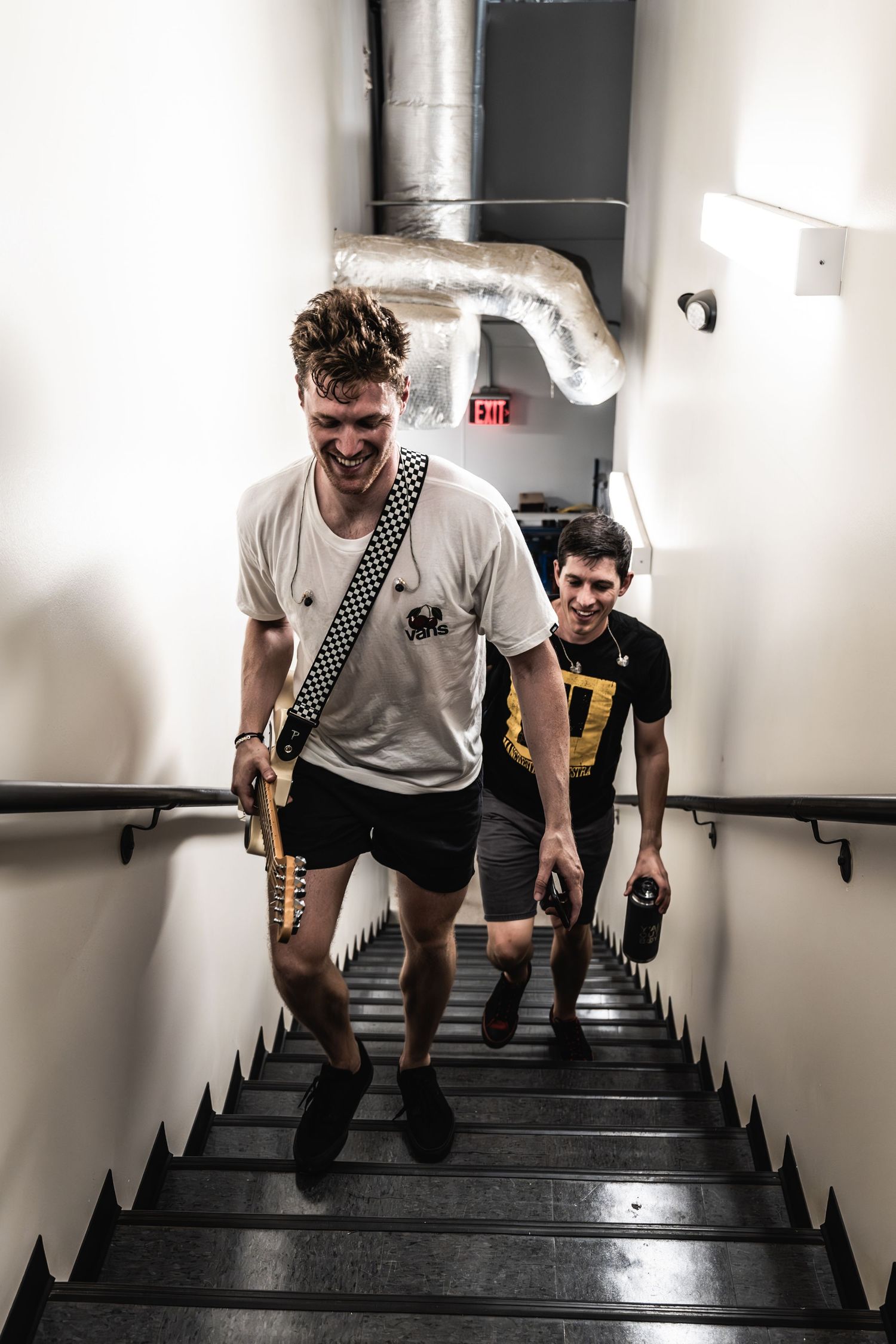 Musicians carrying instruments up stairs in a white-walled venue corridor.