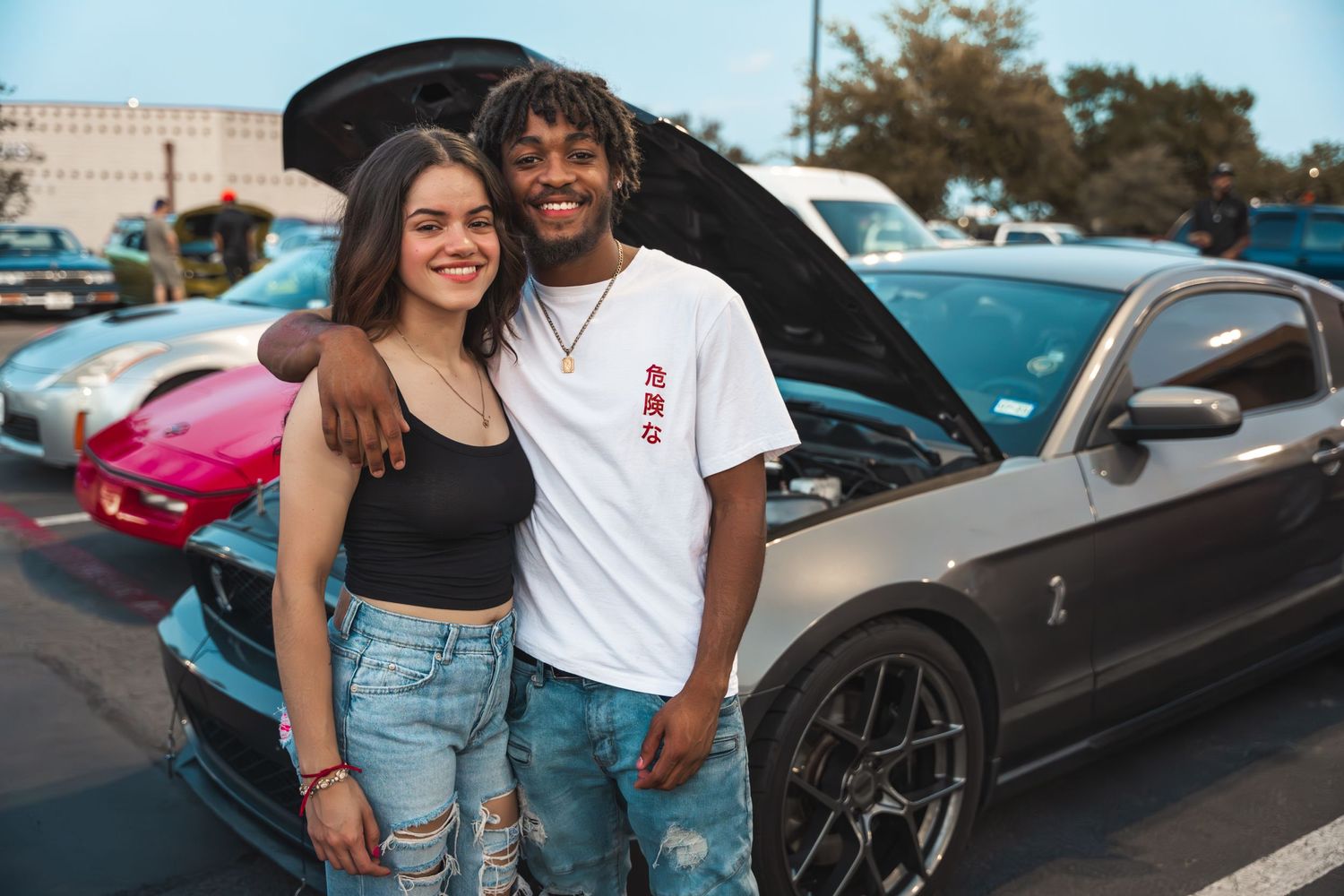Car enthusiasts pose in casual attire at an outdoor auto meet gathering.