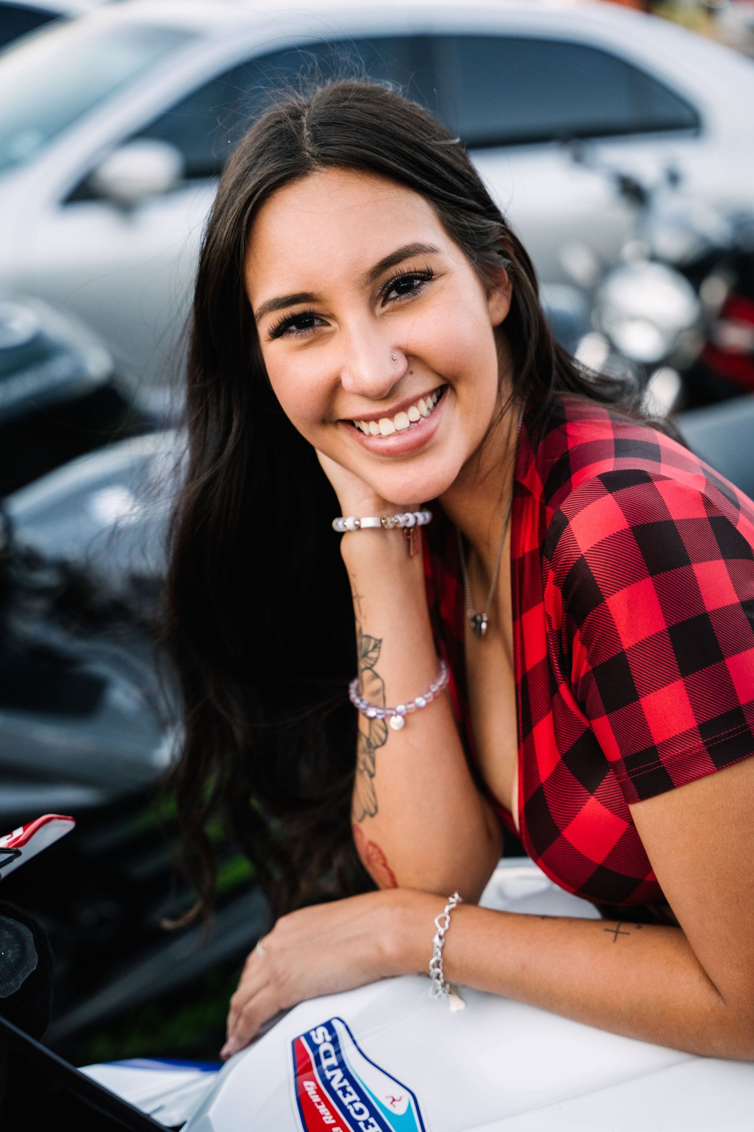 A model in a red buffalo plaid shirt and jewelry poses by cars at an outdoor event.