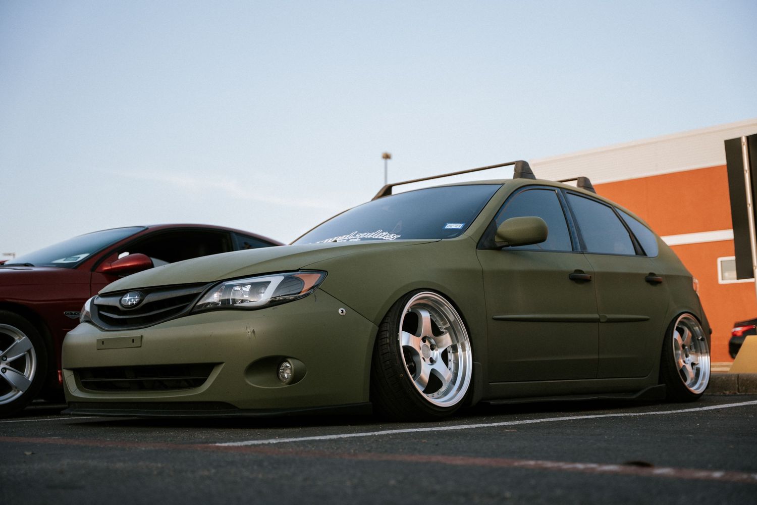 Lowered army green Subaru Impreza with aftermarket wheels parked against orange building.