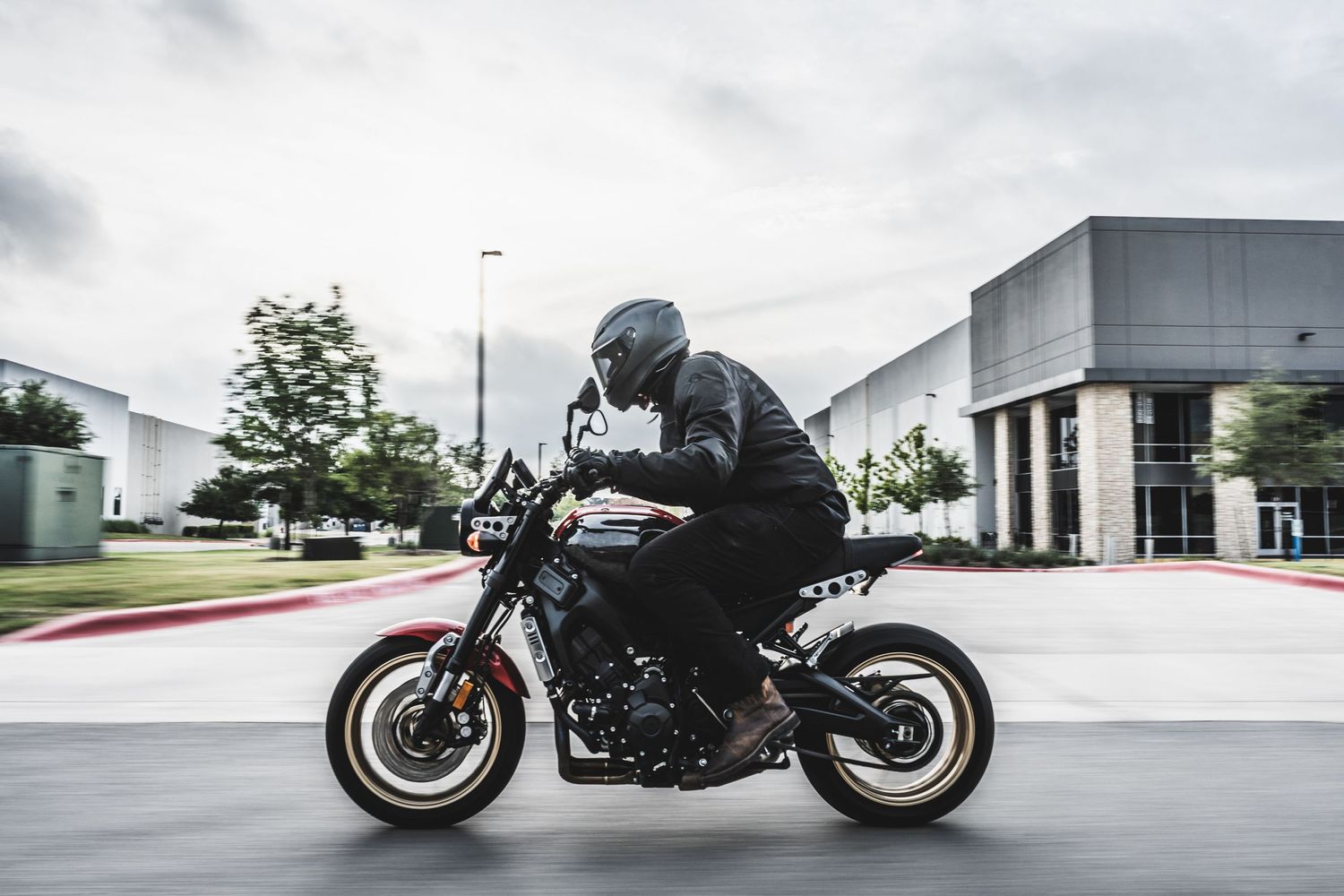 A motorcyclist in black gear rides a modern motorcycle in an urban setting on an overcast day.