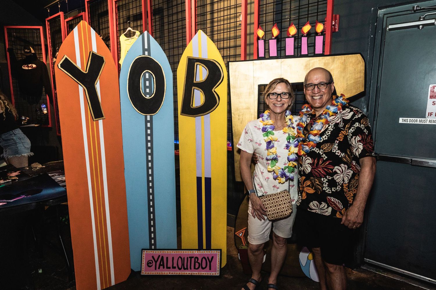 Two people posing in front of colorful surfboard-style wall display with YallOutBoy signage.