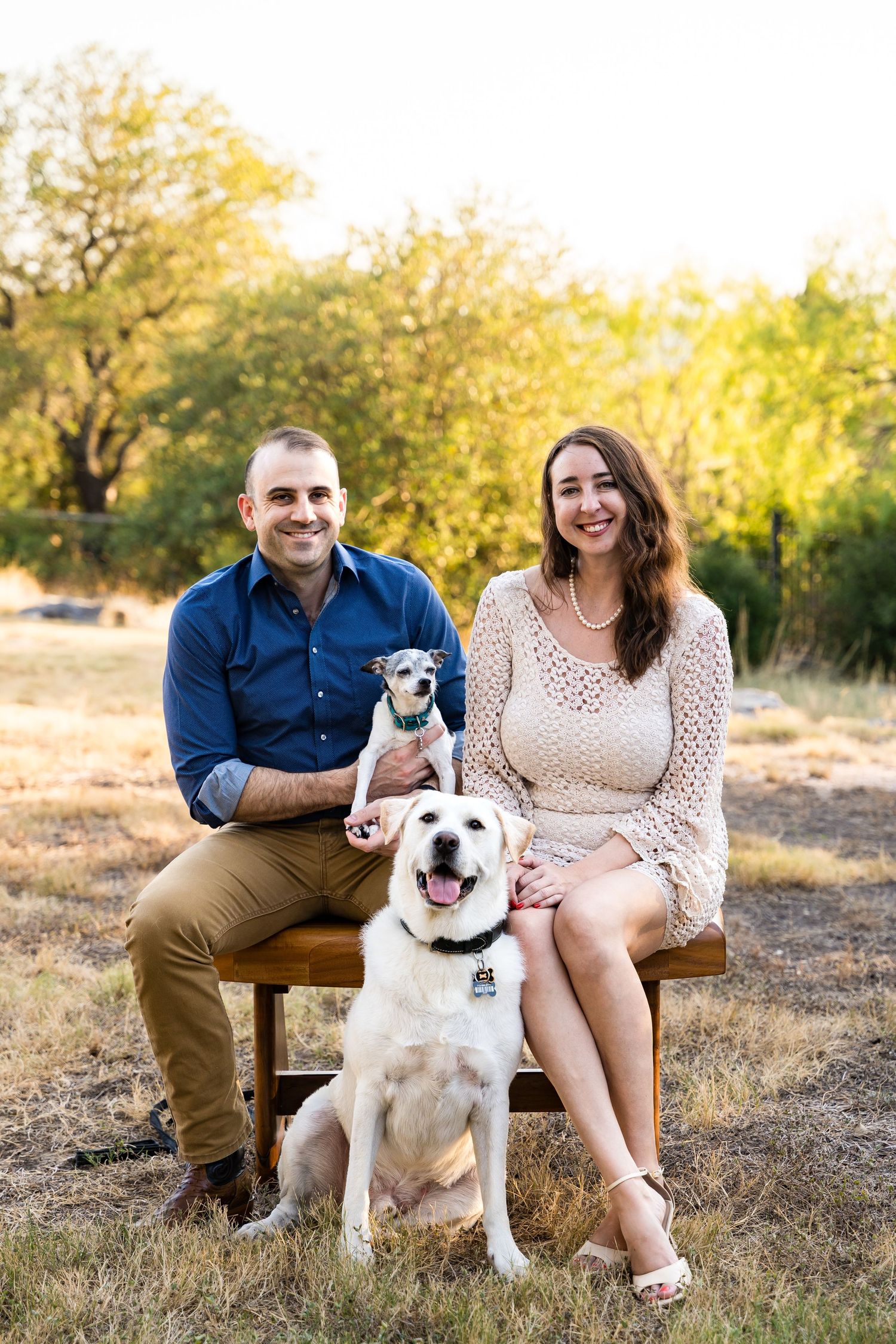 Couple seated outdoors with two dogs against natural landscape.