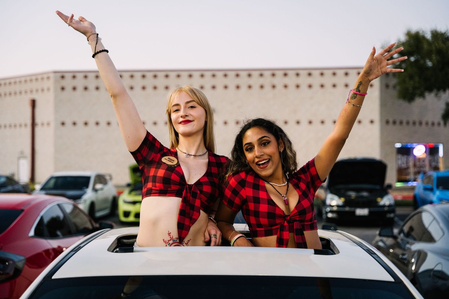 Friends in matching red plaid tops pose joyfully through car sunroof at evening gathering.