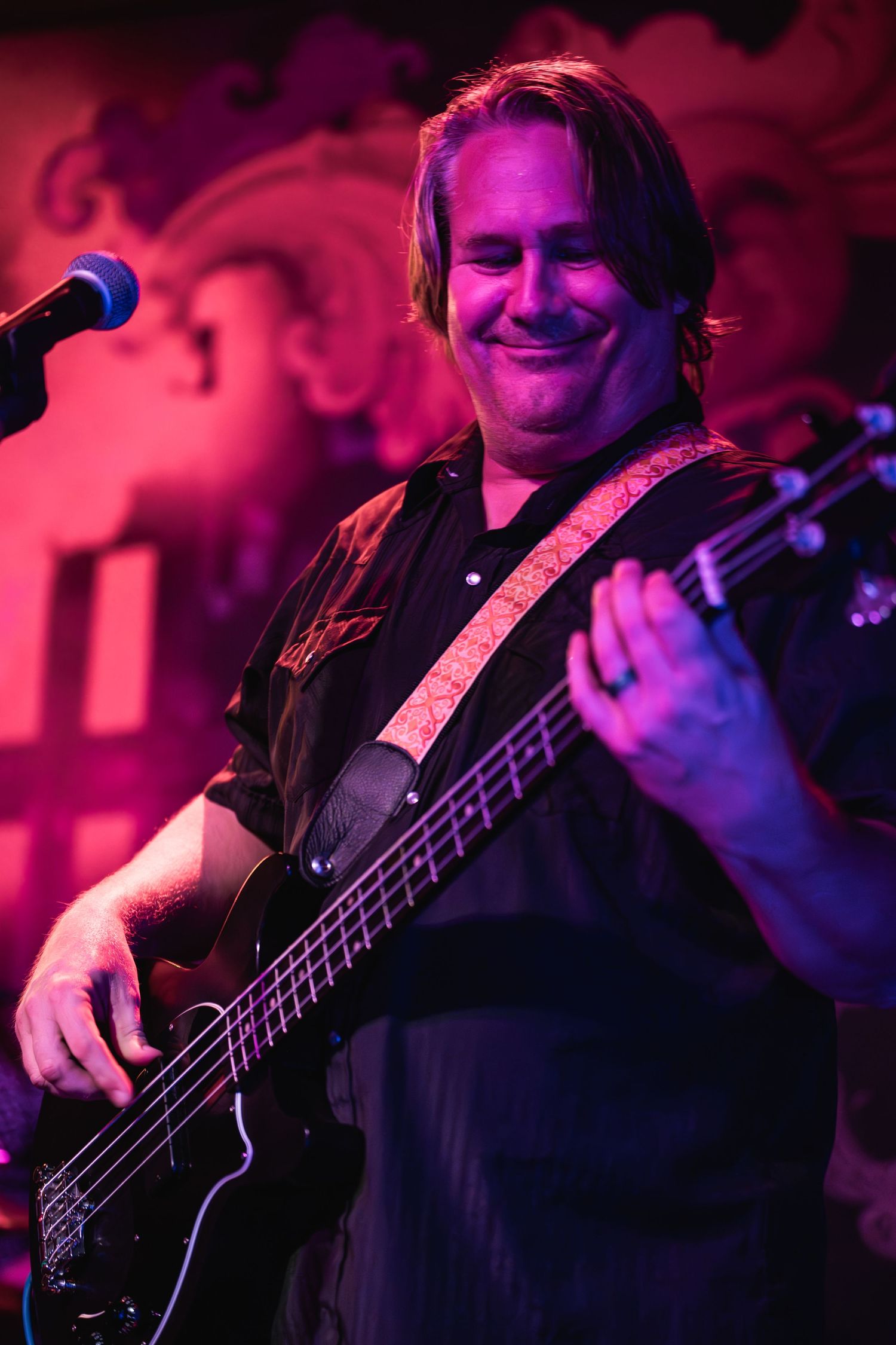 Bass player smiles while performing under pink stage lighting with ornate backdrop.