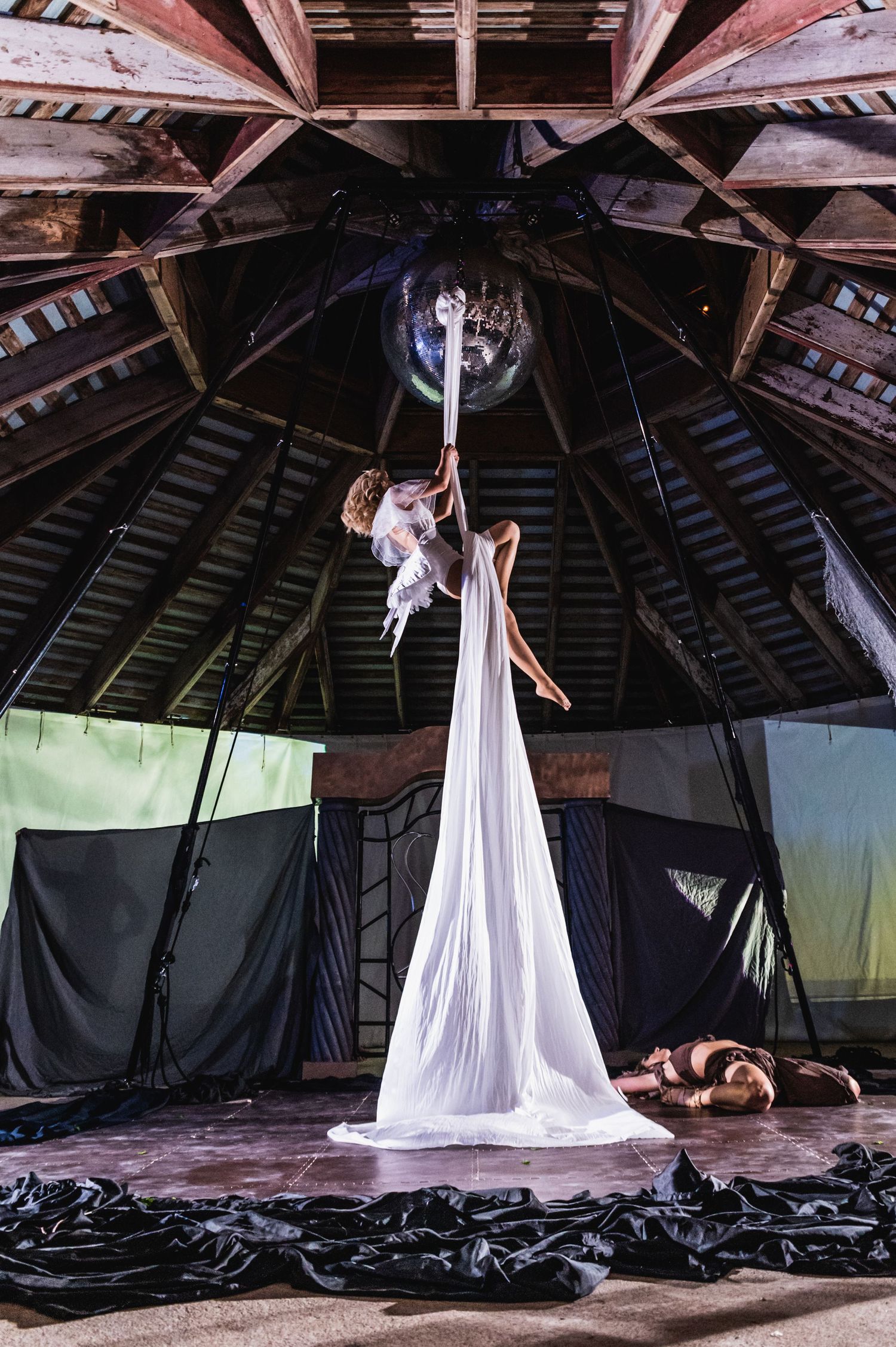 An aerial silk performer in white costume suspended from a wooden domed ceiling.