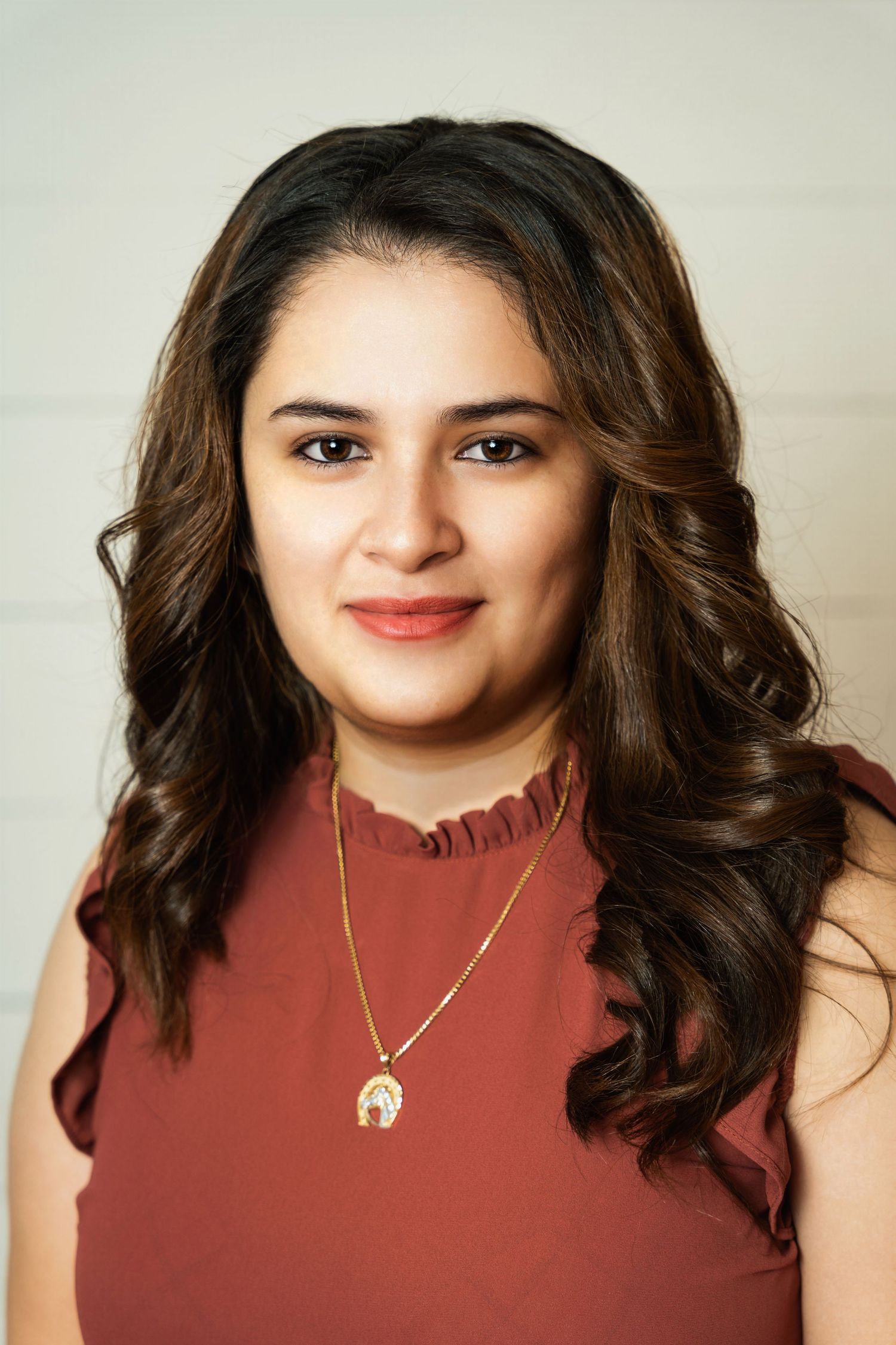 Professional portrait of person wearing burgundy sleeveless top with gold necklace against light background.