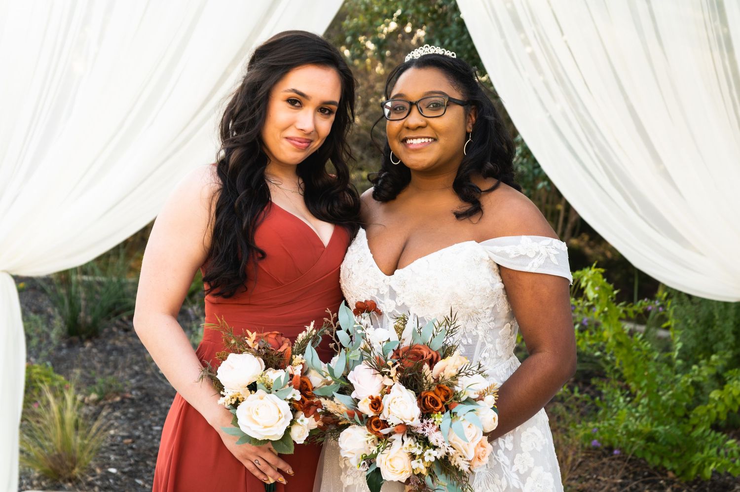 Bride and bridesmaid holding white and rust-colored bouquets pose under white draping at outdoor wedding.