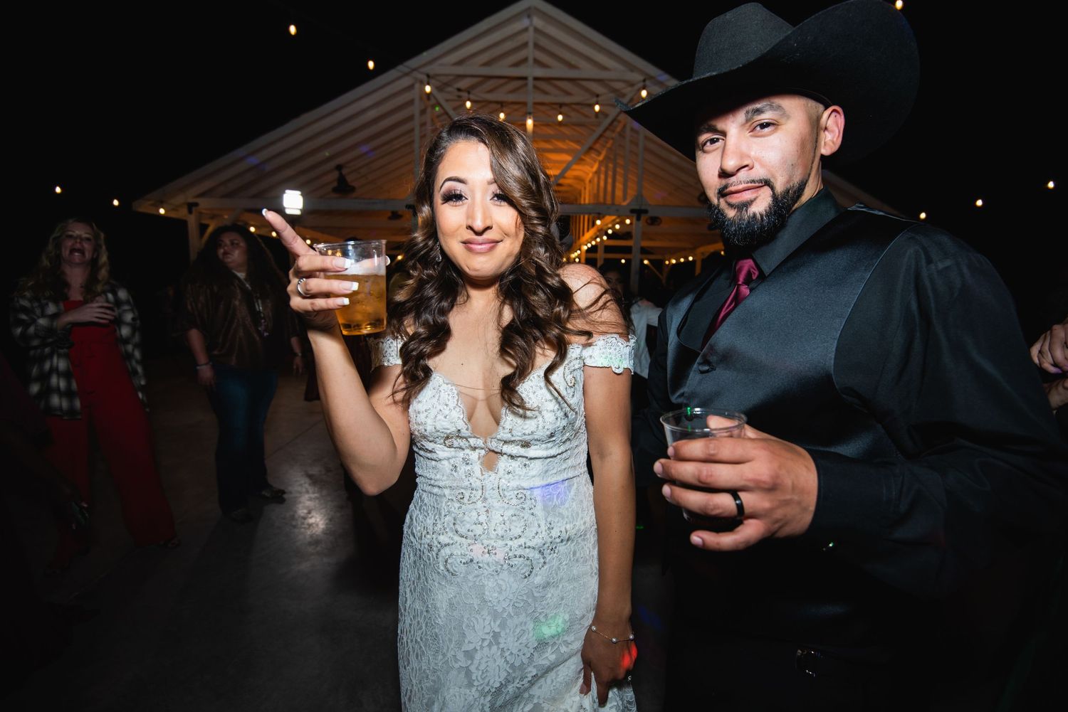 A couple toasts with drinks at their wedding reception under twinkling lights.
