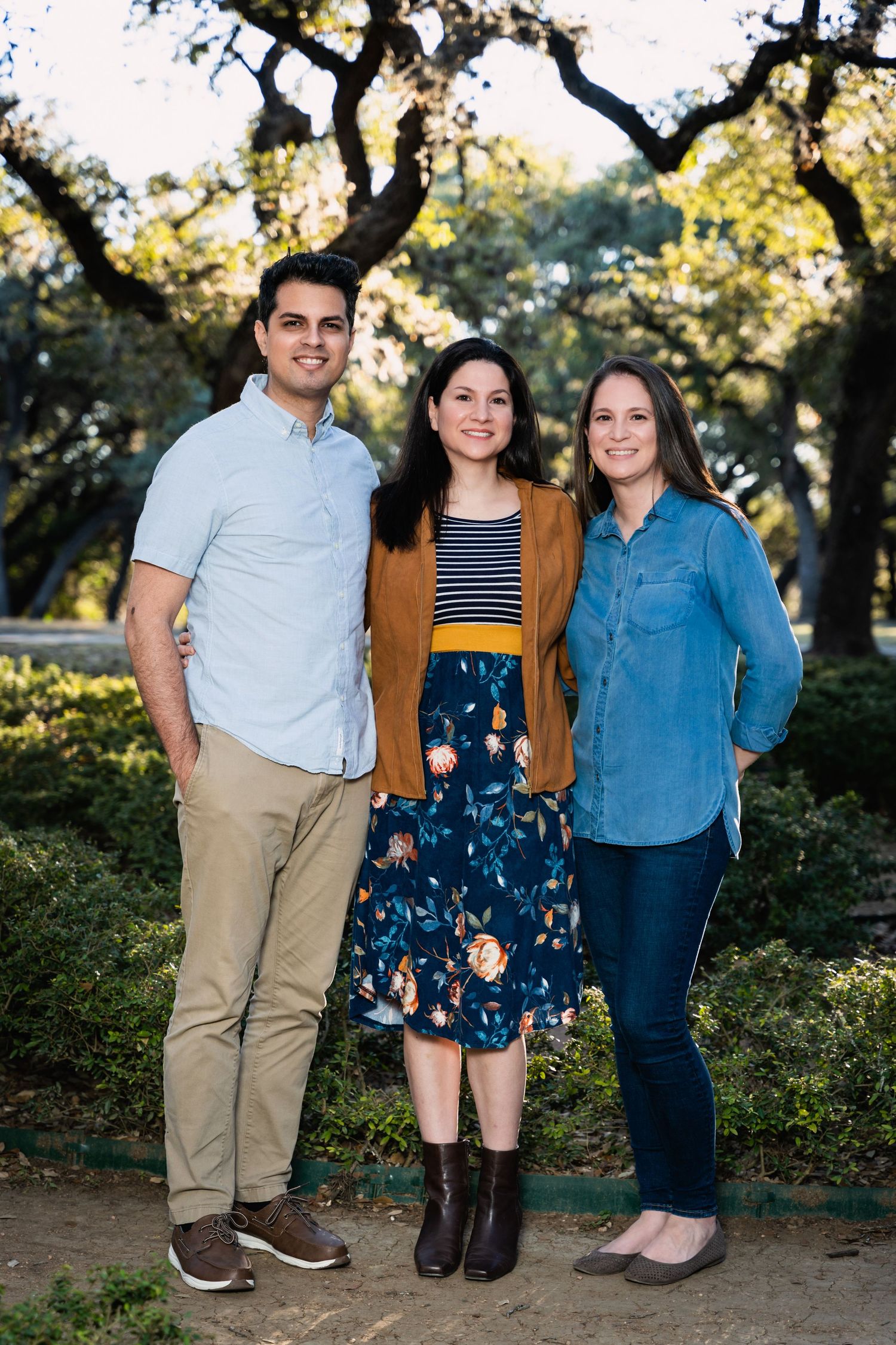 Three people pose together on garden path with trees in background.