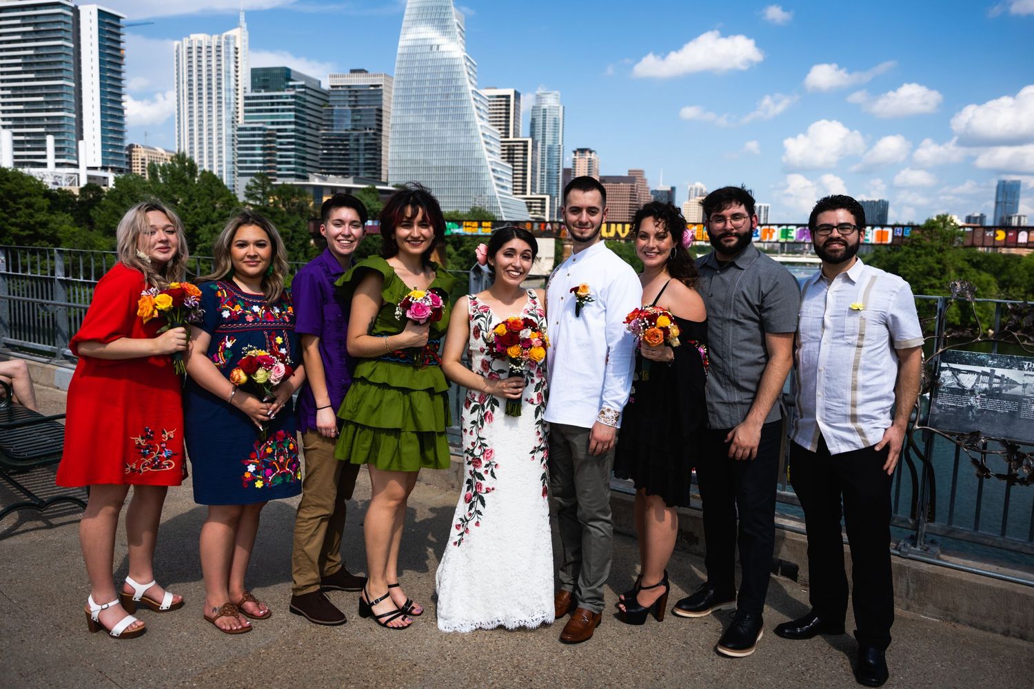 A wedding party poses on an outdoor deck with a city skyline visible in the background.