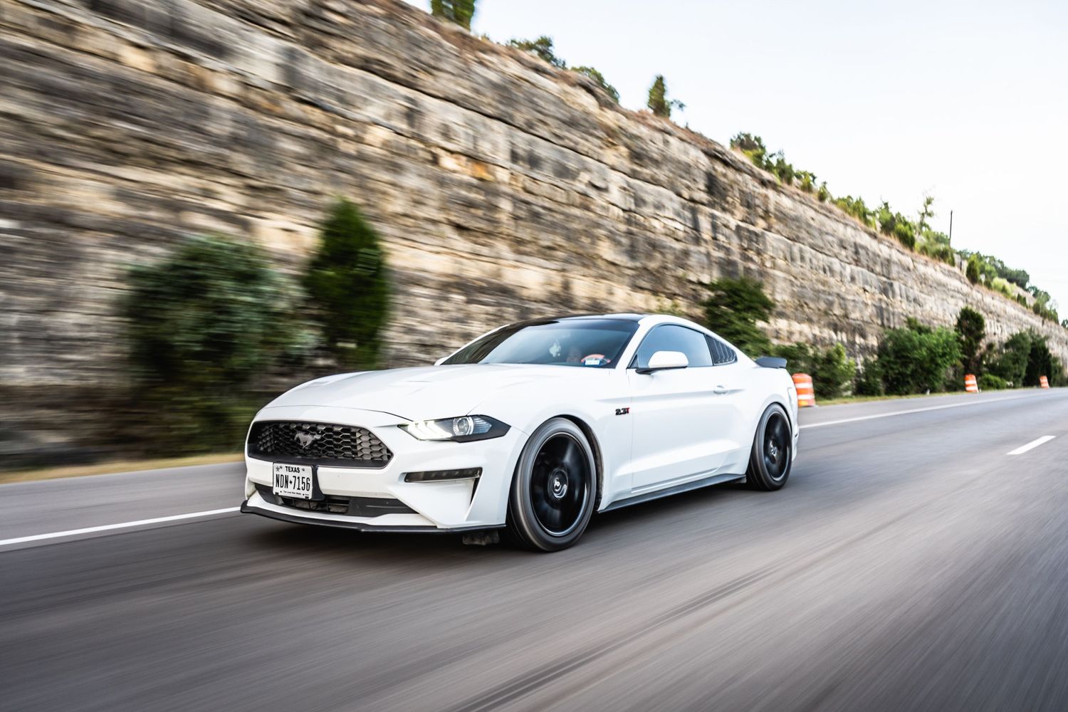 White Ford Mustang with black wheels photographed speeding past a rock wall.