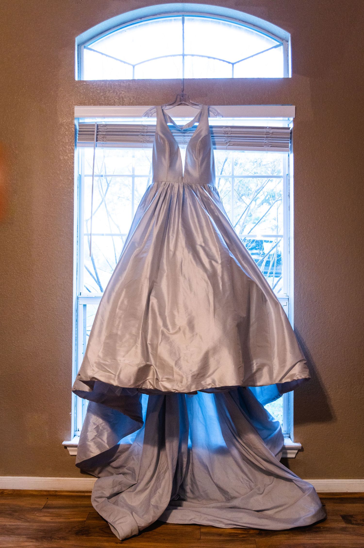 A silver ballgown wedding dress hangs elegantly in front of a window with natural light.