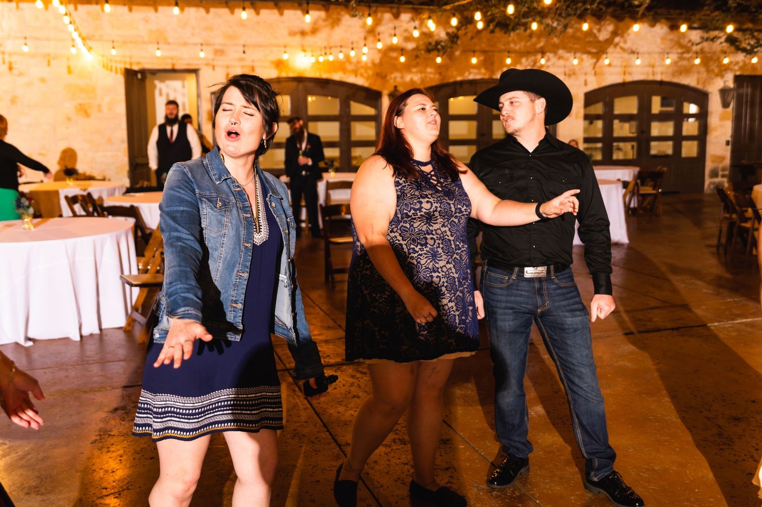 Party guests dance enthusiastically at a wedding reception with string lights in the background.