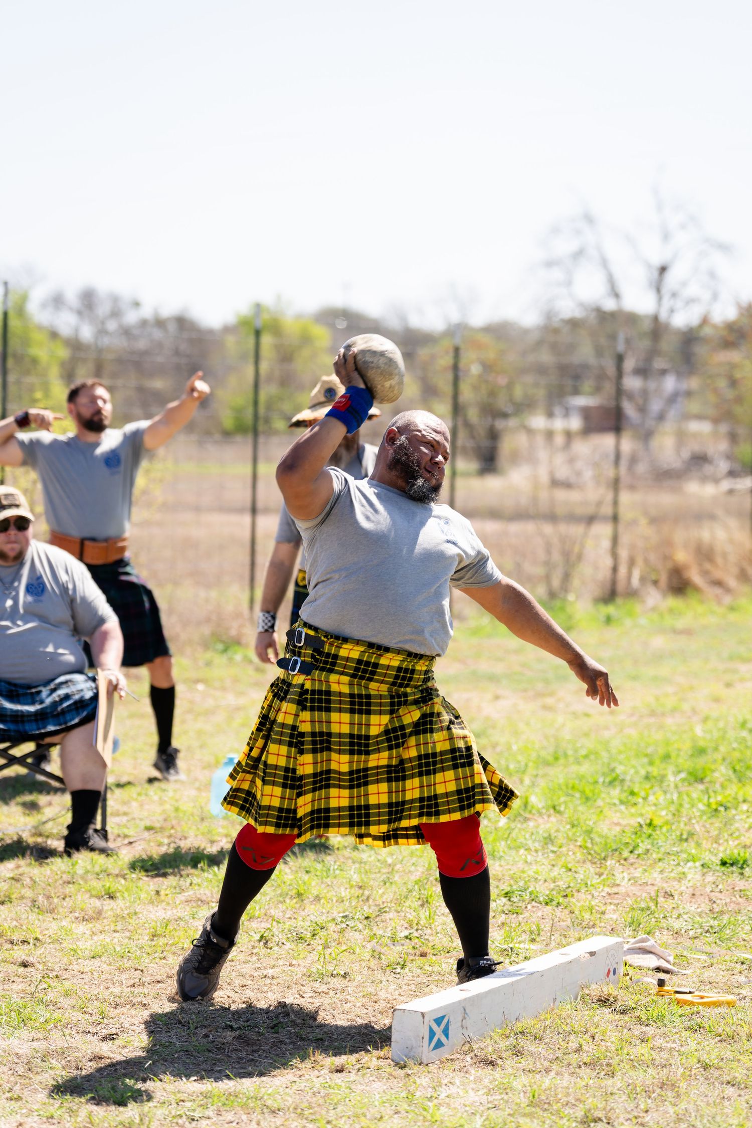 Man in yellow tartan kilt throws stone at Highland Games competition outdoors.