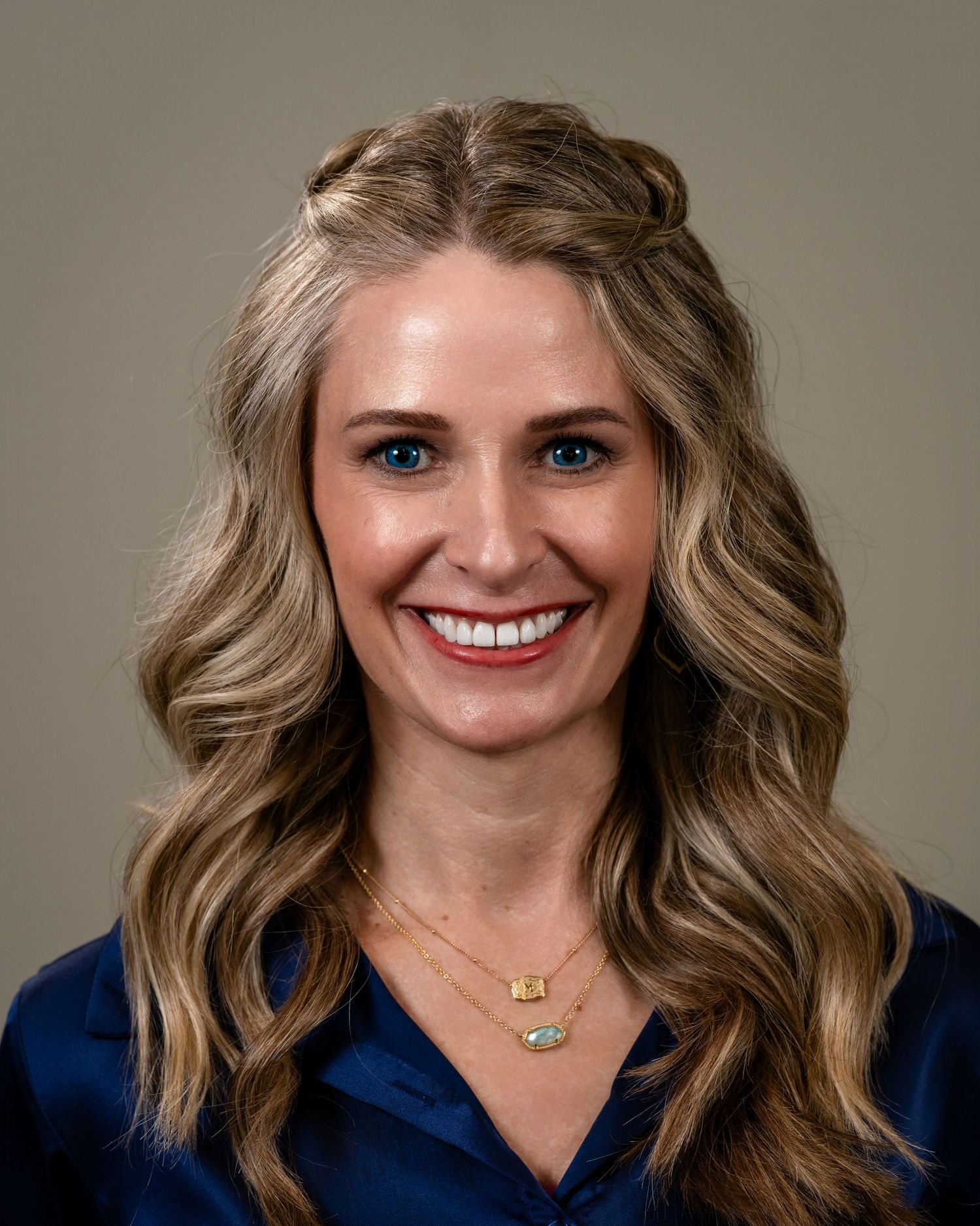 Professional headshot of a smiling woman with long blonde wavy hair wearing a navy blue top and gold necklace.